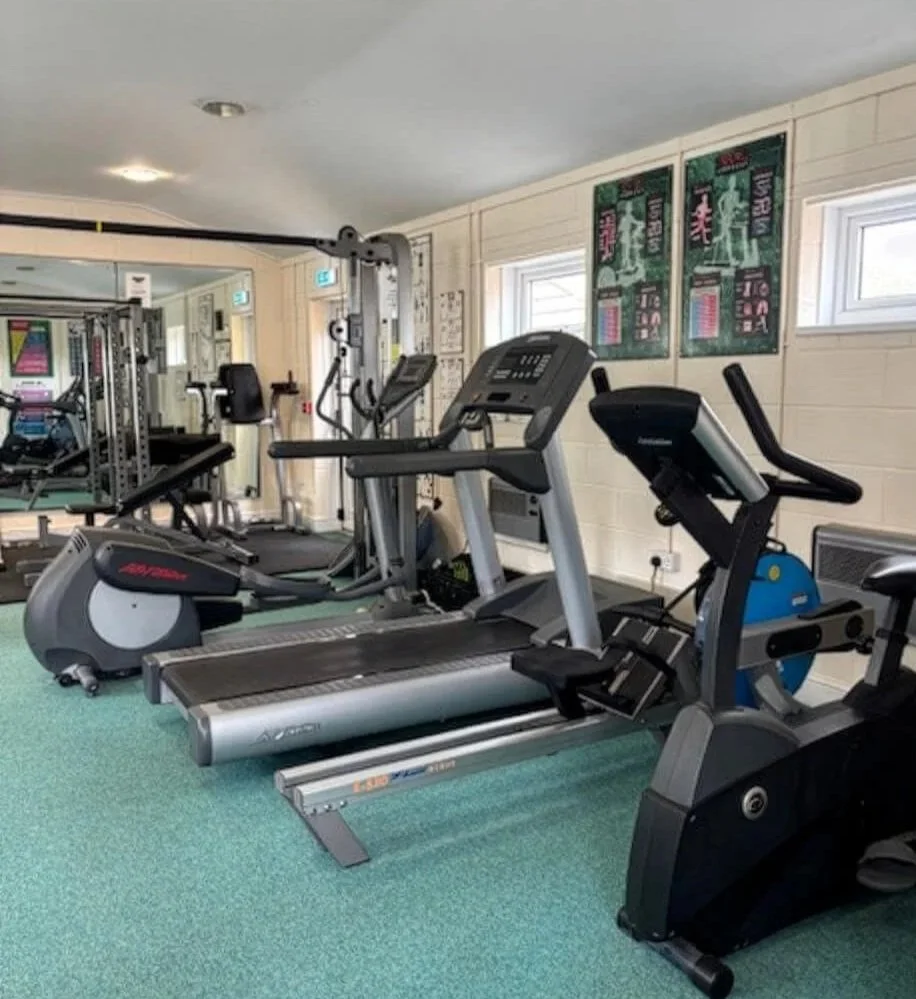 A gym room with exercise equipment including a treadmill, an elliptical machine, and a recumbent bike, with mirrors and posters on the walls.