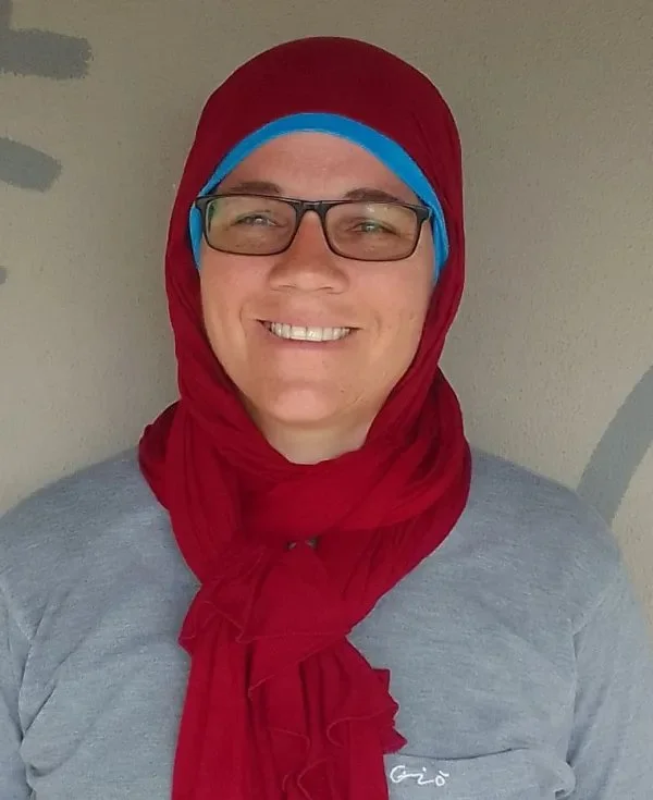 Person wearing glasses and a red headscarf tied around the neck, smiling, with a light-colored background.