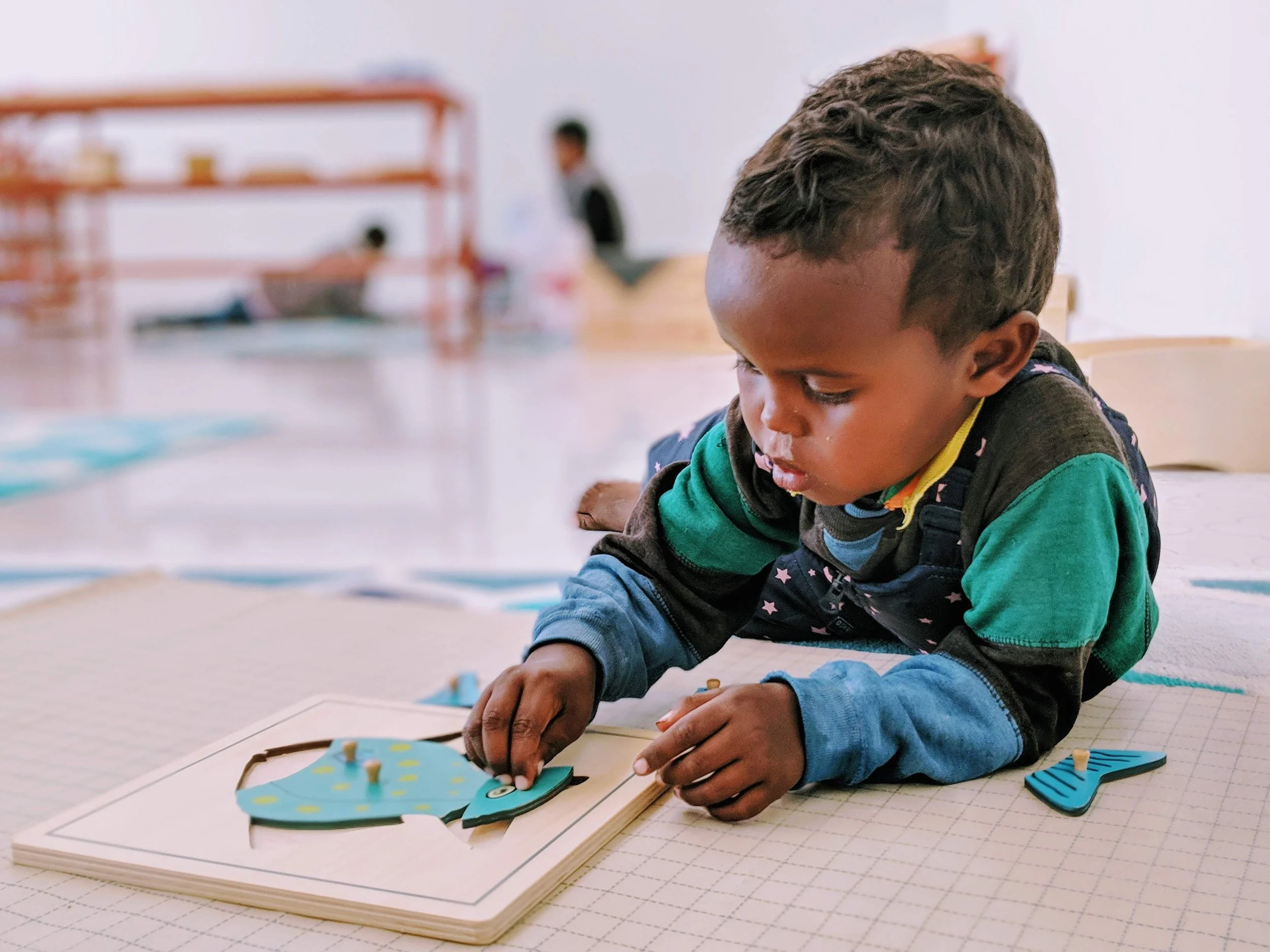 A young boy student lying on a floor playing with a Montessori fish-shaped puzzle in a classroom setting.