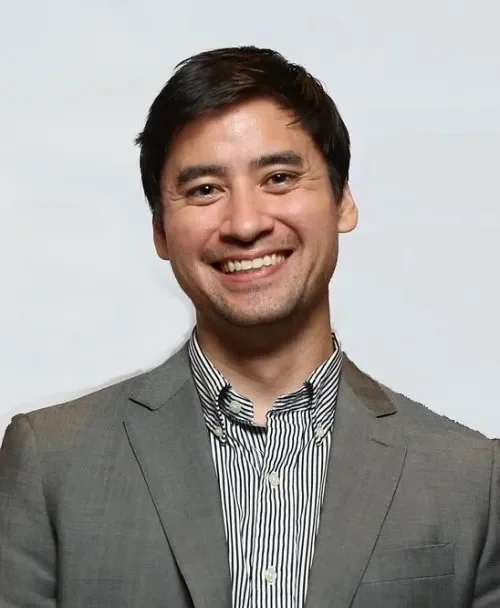 A smiling man in a gray suit and striped shirt standing in front of a light background.