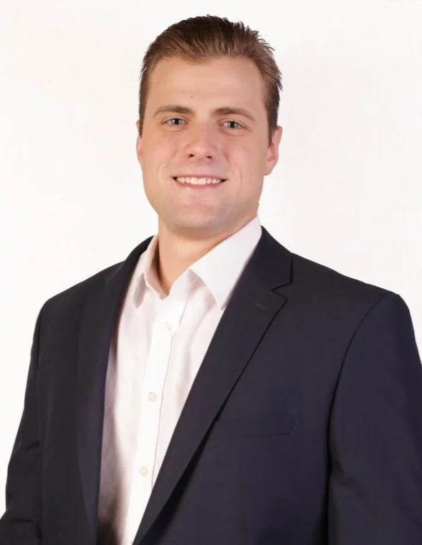 A young man with short light brown hair, wearing a white dress shirt and a black suit jacket, smiling against a plain white background.