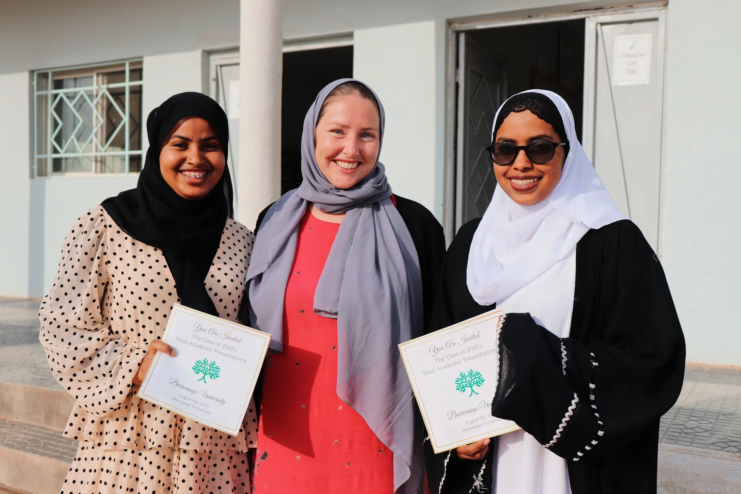 Three women, two wearing hijabs and holding diplomas, standing outside a building and smiling.