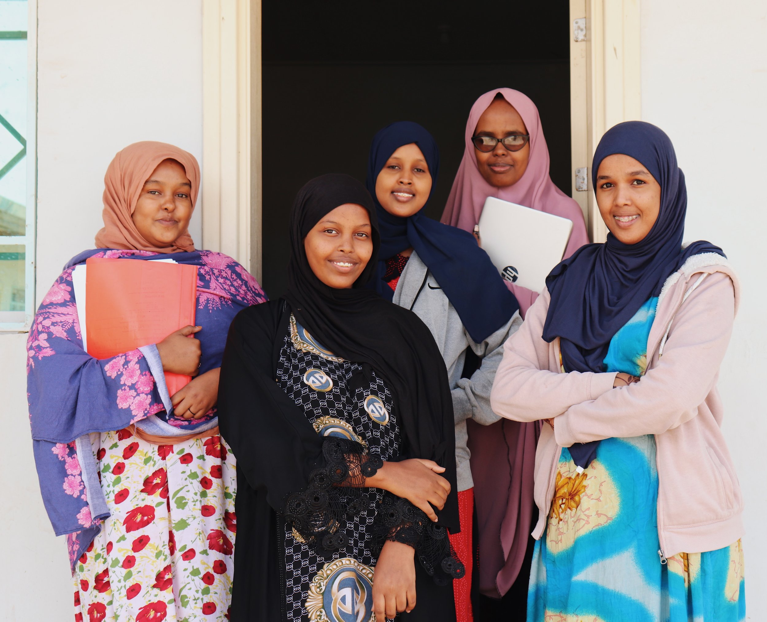 Group of five women wearing hijabs and colorful dresses standing in front of a doorway, smiling at the camera.