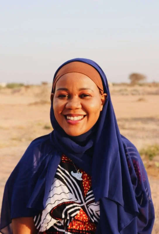 Smiling woman wearing a brightly colored, patterned dress and a dark blue hijab, standing outdoors in a desert landscape with a clear sky.
