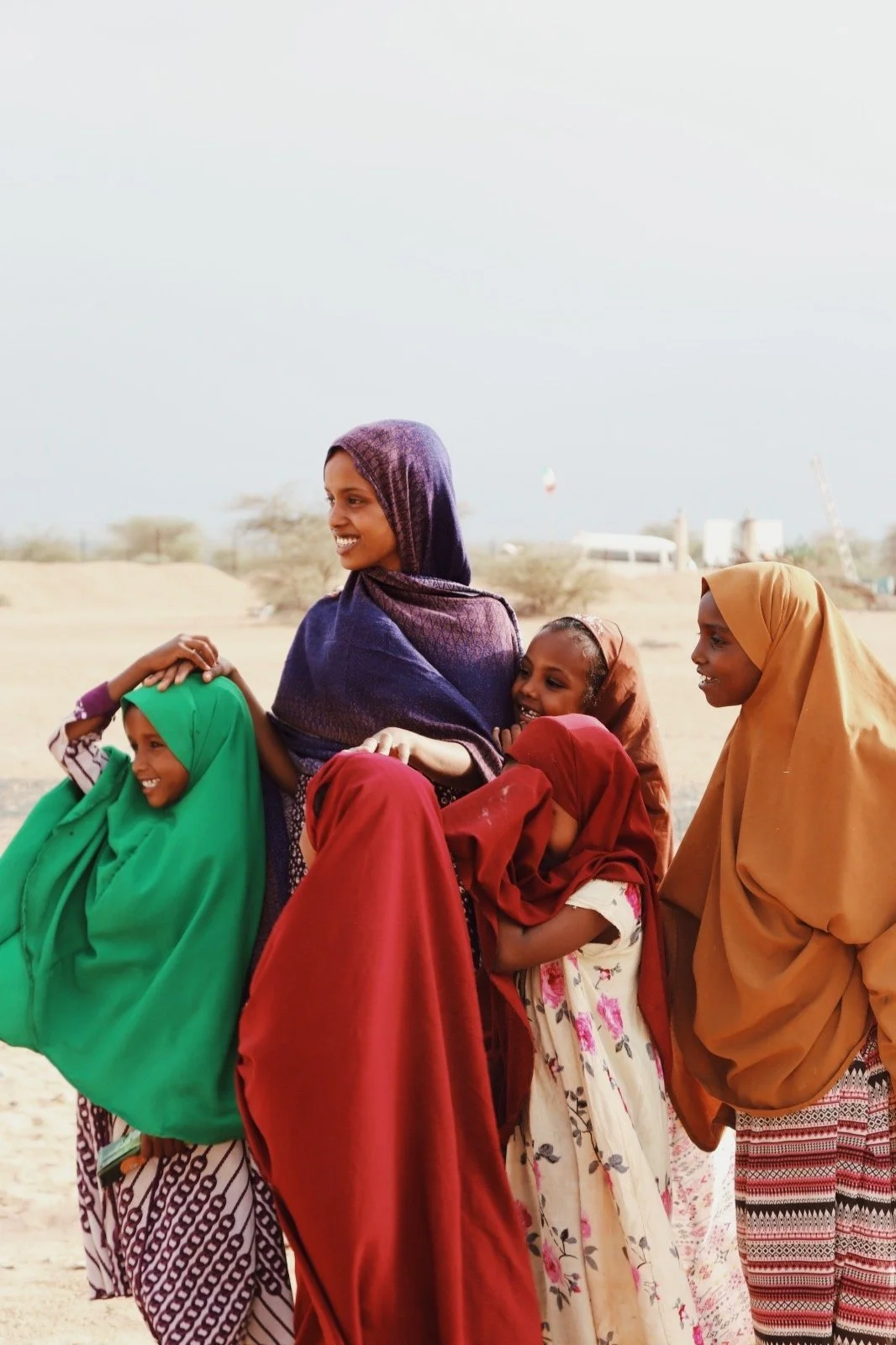 Group of women and children in colorful traditional clothing smiling and got in conversation outdoors in a desert landscape.