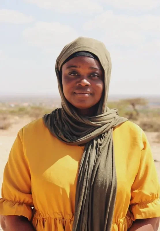 A woman in a yellow dress and a khaki headscarf standing outdoors in a desert landscape with sparse vegetation and a cloudy sky.