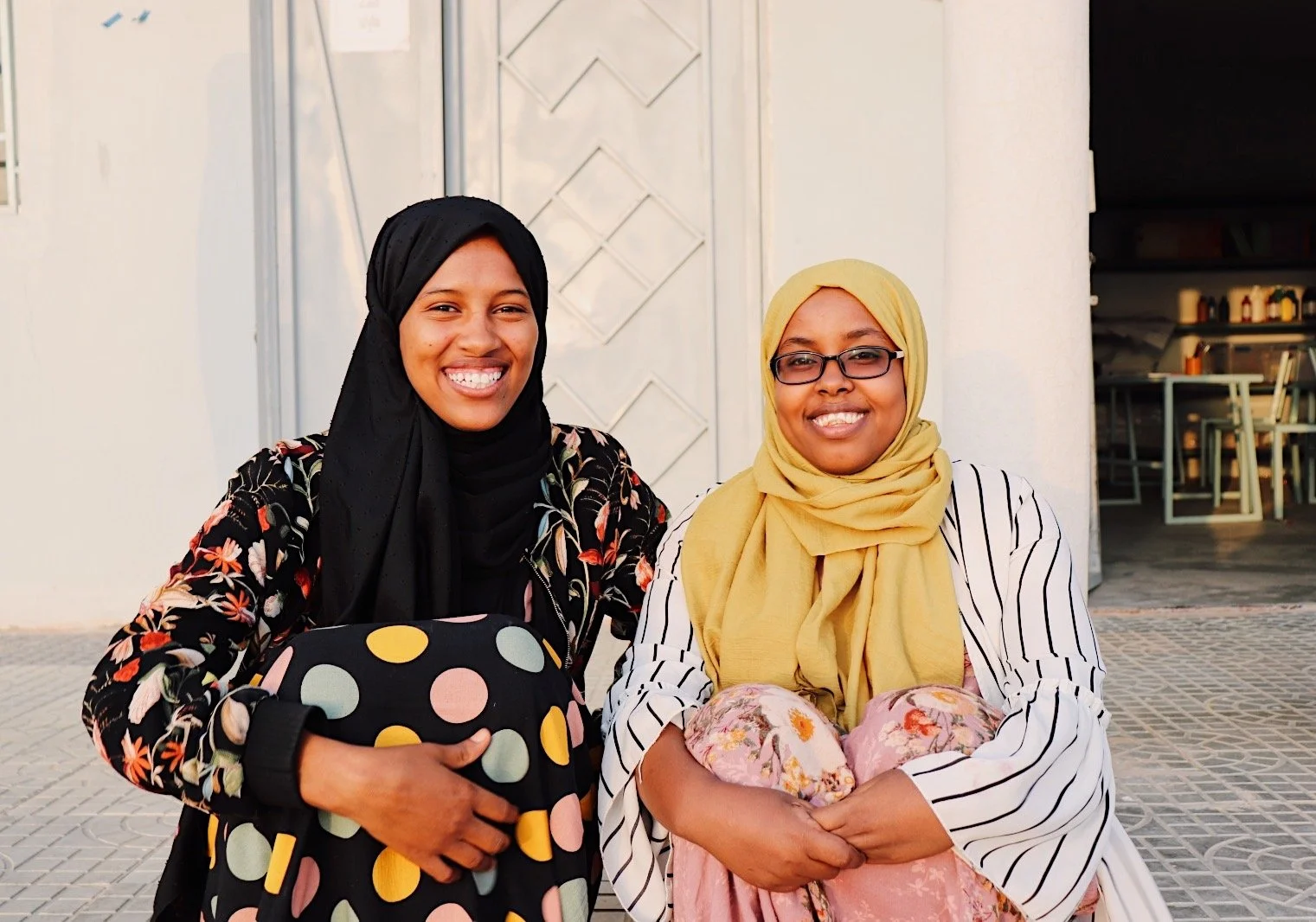 Two smiling women sitting outside, one wearing a black hijab and floral blazer holding a colorful polka dot bag, the other wearing glasses, a yellow hijab, and a striped blazer, holding a floral pillow.
