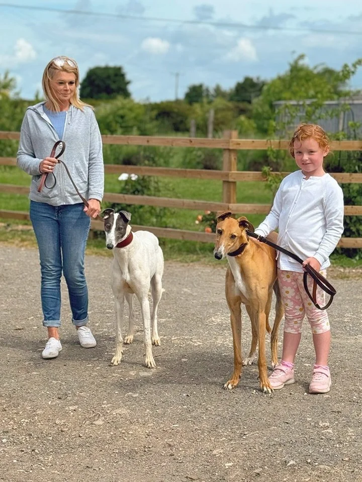 A woman and a young girl each holding a dog on a leash in an outdoor setting with a wooden fence and trees in the background.