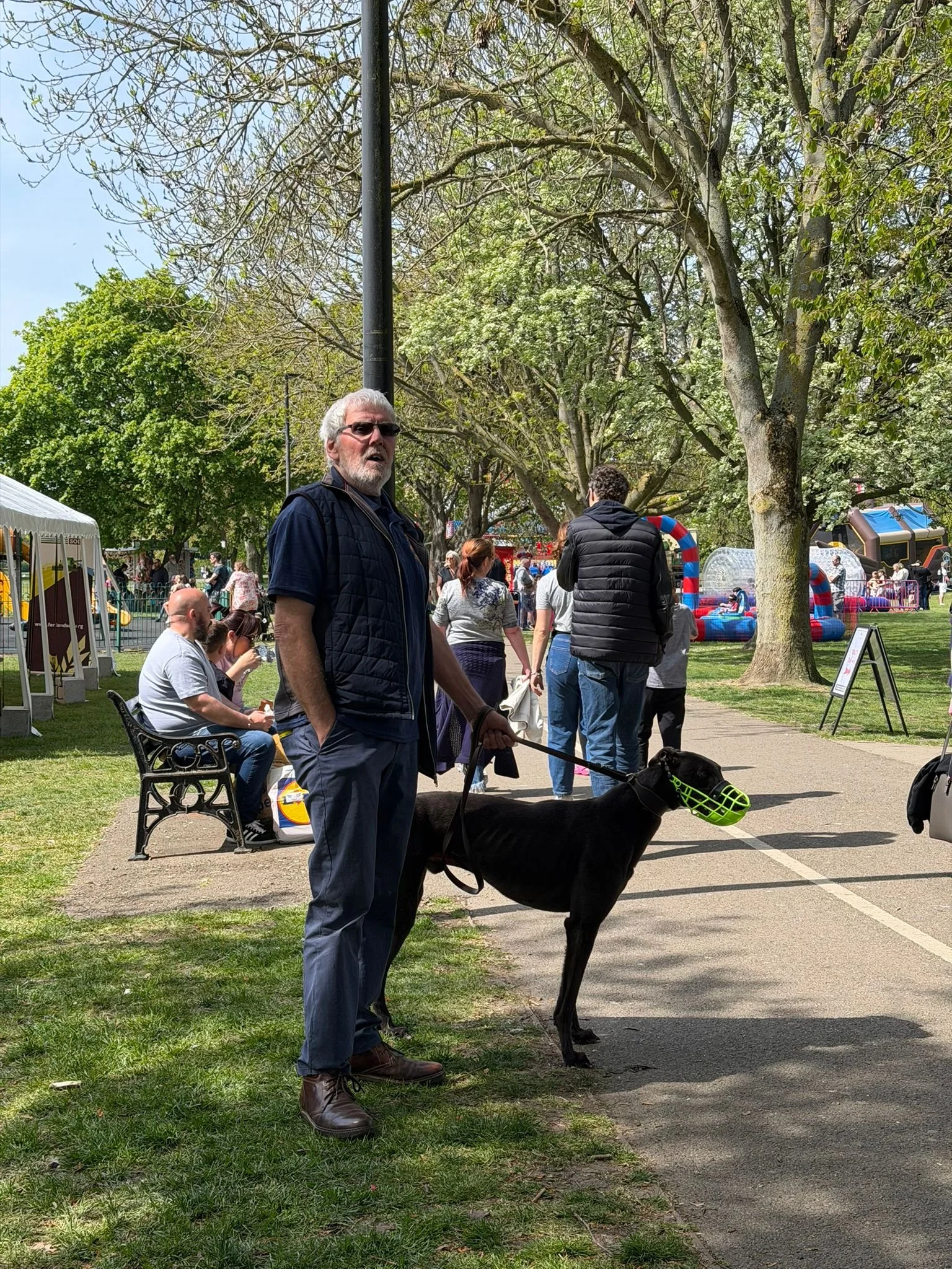 A man with gray hair and a beard wearing sunglasses, a dark vest, and blue pants is walking a large black dog wearing a muzzle at an outdoor fair or park on a sunny day. There are other people, trees, and amusement rides in the background.