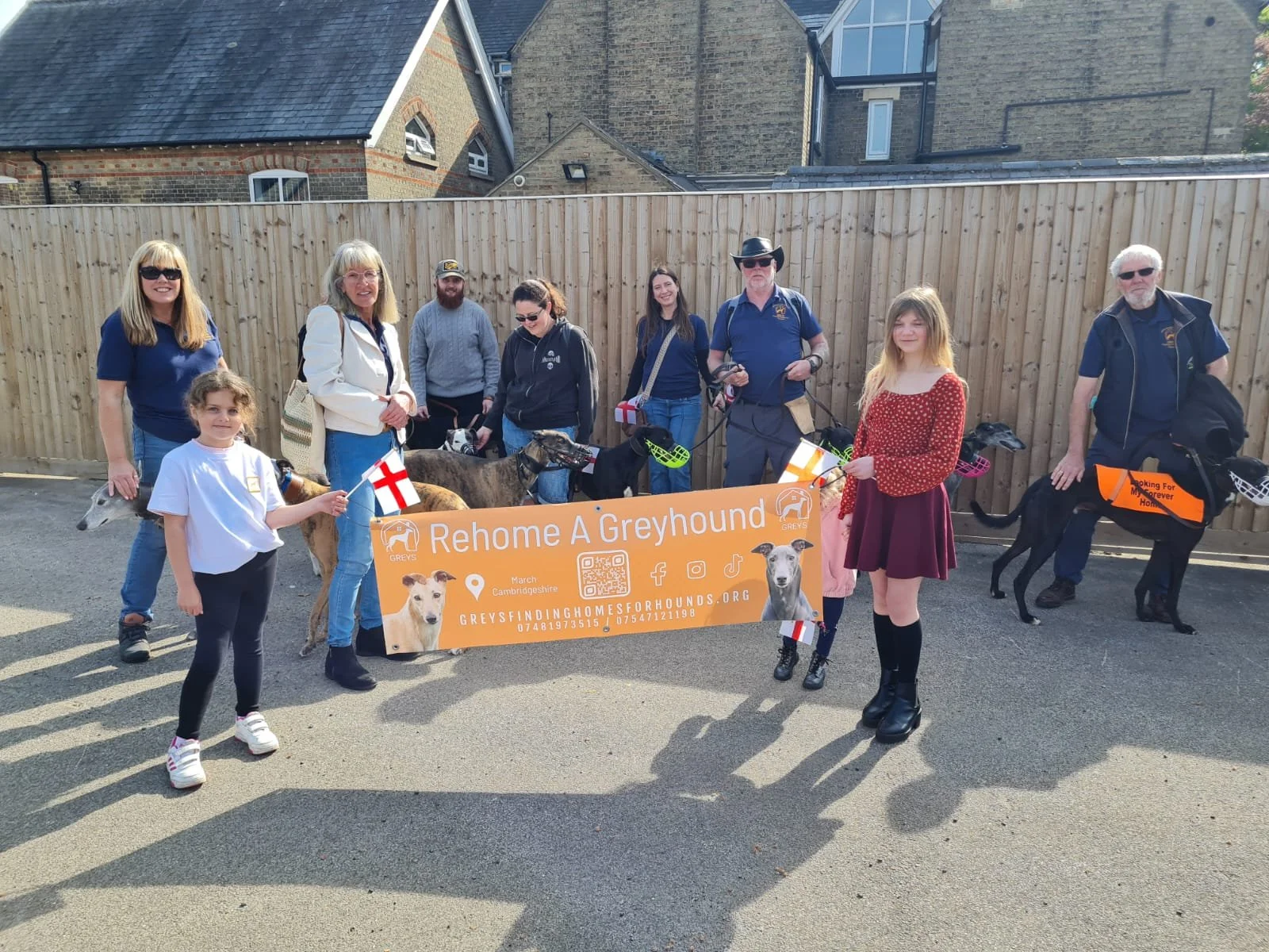 Group of people and dogs participating in a greyhound adoption event outdoors, holding a banner that reads 'Rehome A Greyhound' in front of a wooden fence, with some people holding flags and wearing casual clothing.