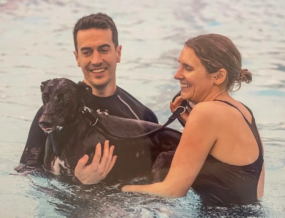A man and a woman happily standing in a body of water, holding a black puppy between them.