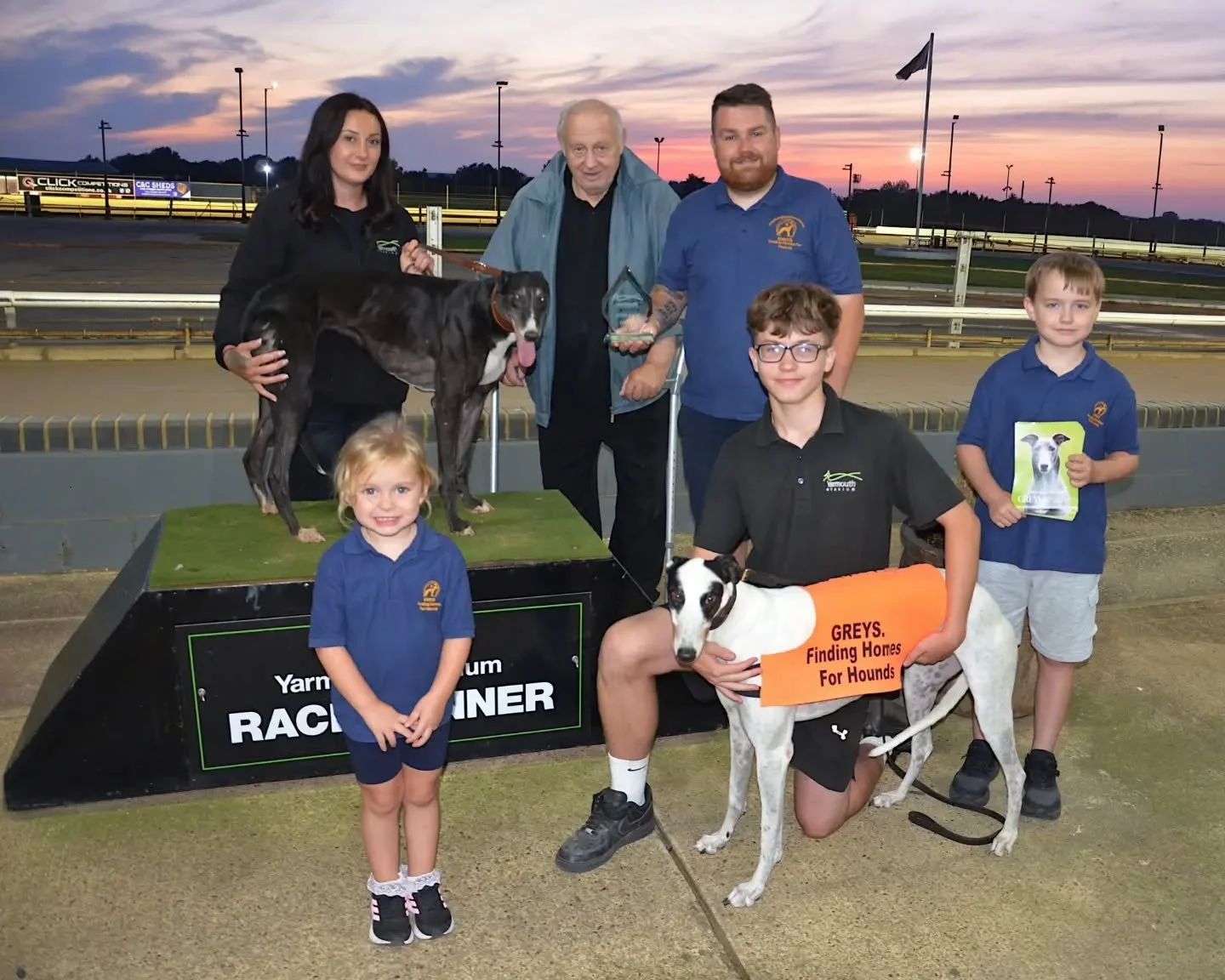Group of children and adults at a racetrack celebrating a greyhound rescue event, with racing dogs on a podium and a sign stating "GREYS. Finding Homes For Hounds."