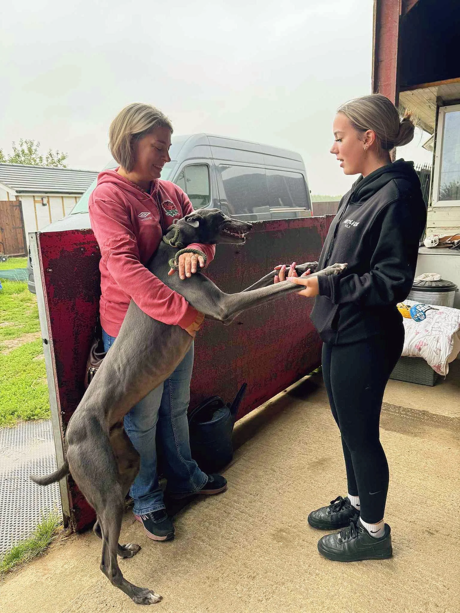 Two women and a greyhound dog standing on its hind legs. One woman is holding the dog, and the other is holding the dog’s front paws. The women are inside a garage with a vehicle and outdoor scenery in the background.
