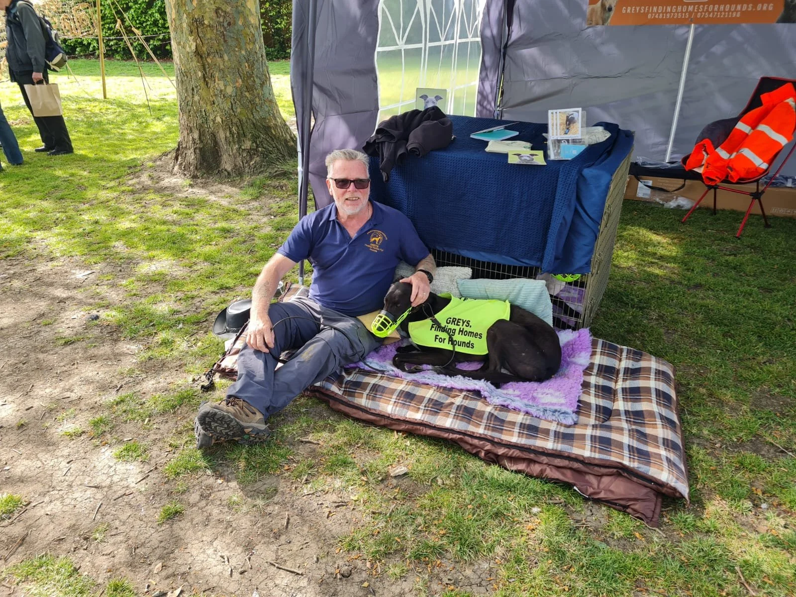A man sitting on a blanket on the grass with a black dog wearing a fitness vest, under a canopy at an outdoor event. The man is wearing sunglasses and a blue shirt, smiling, with some informational materials on a table behind him.