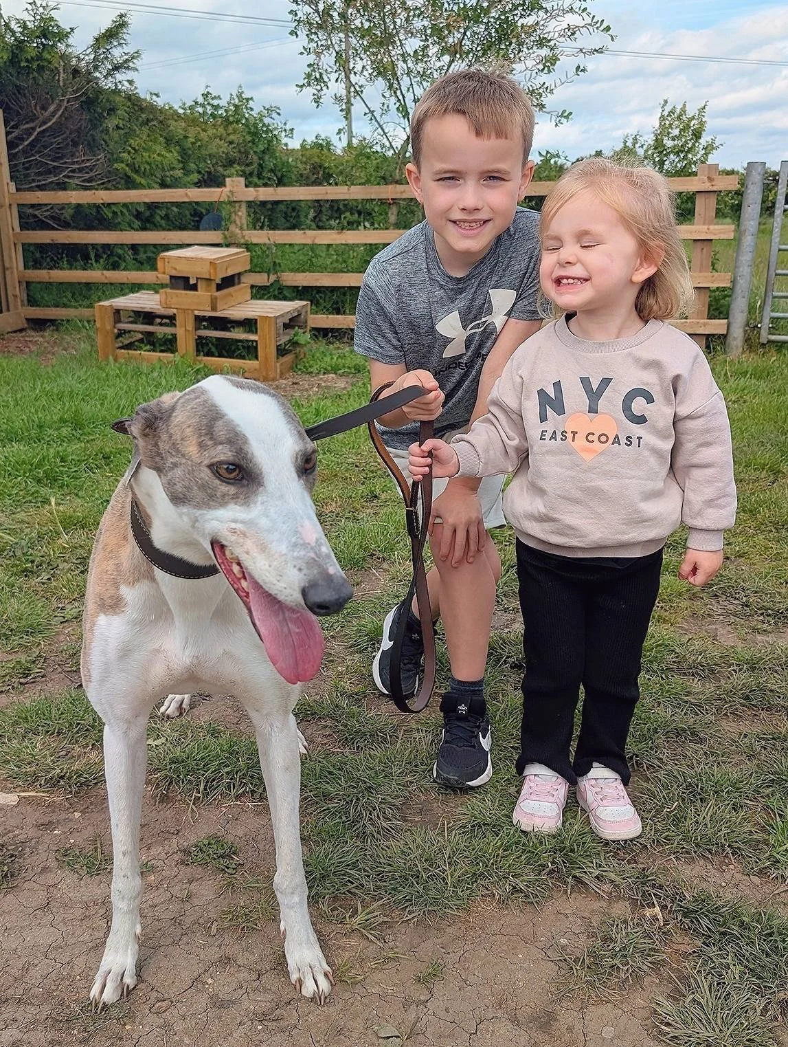 A boy and a girl smiling with a dog in a fenced yard on a cloudy day.