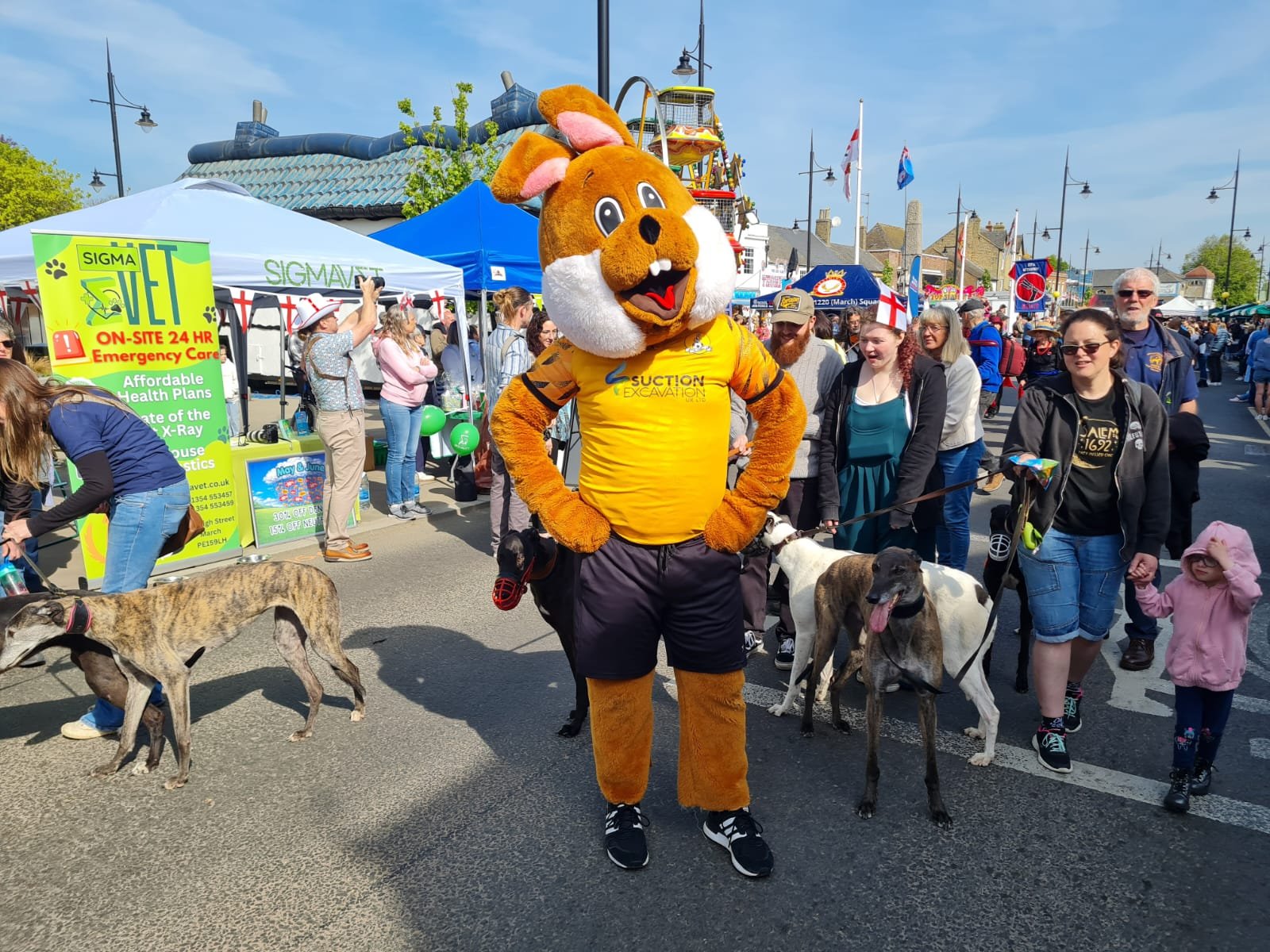 Person in a dog costume at a crowded outdoor event with tents and people, some with dogs, under a partly cloudy sky.