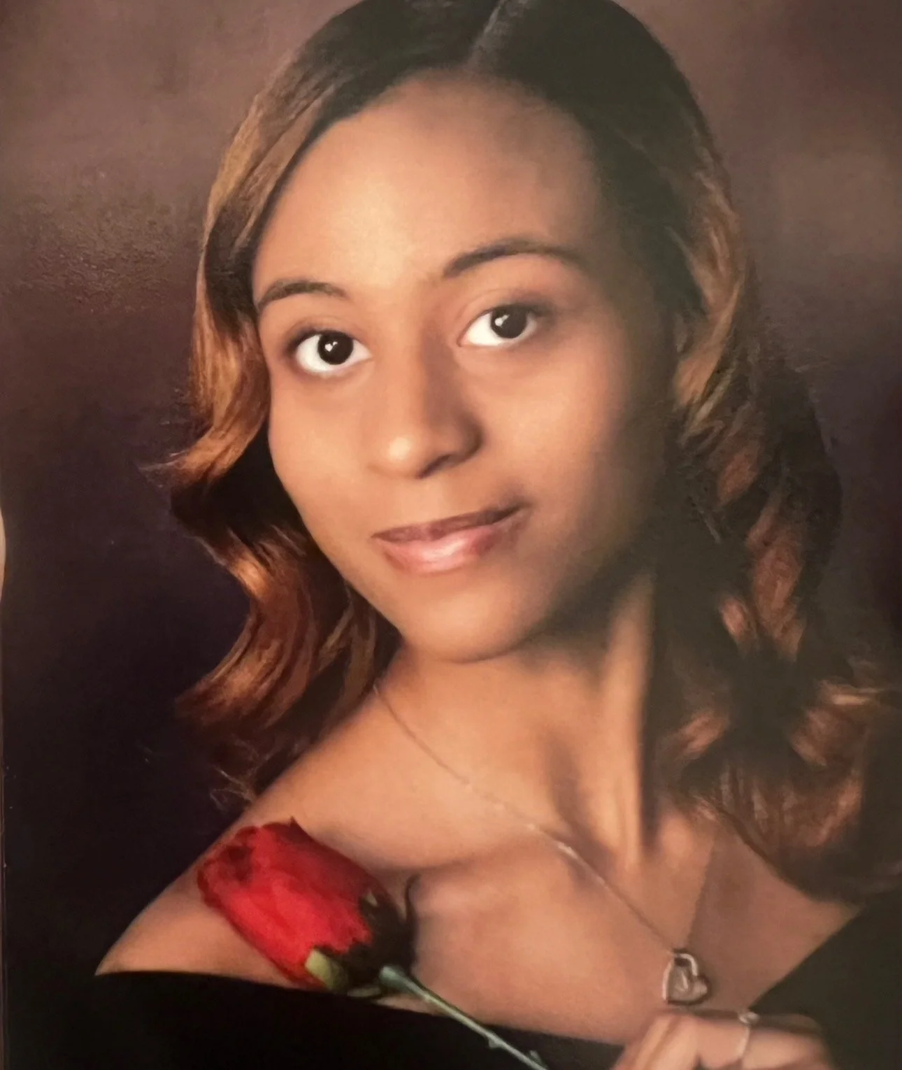 Portrait of a young woman with shoulder-length wavy hair, wearing a red dress with floral patterns and a heart-shaped necklace, against a dark background.