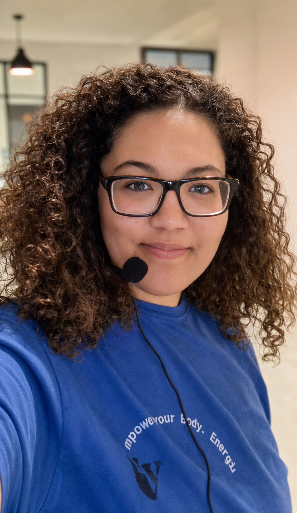 A woman with curly brown hair wearing glasses and a blue shirt, smiling at the camera, with a headset microphone on