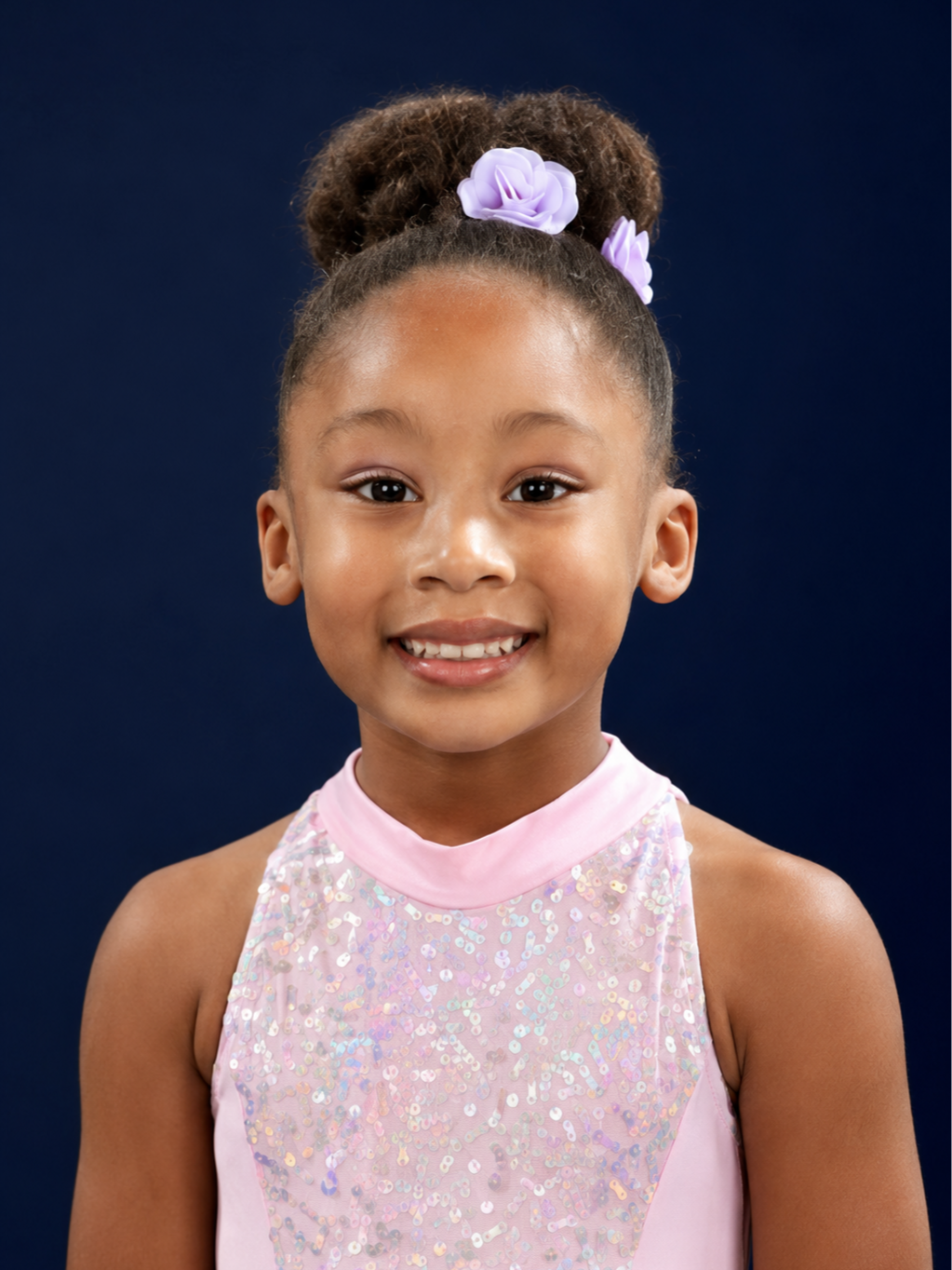 Young girl with curly hair styled in a bun with purple flower hair accessories, wearing a light pink dress with iridescent sequins, smiling against a dark blue background.
