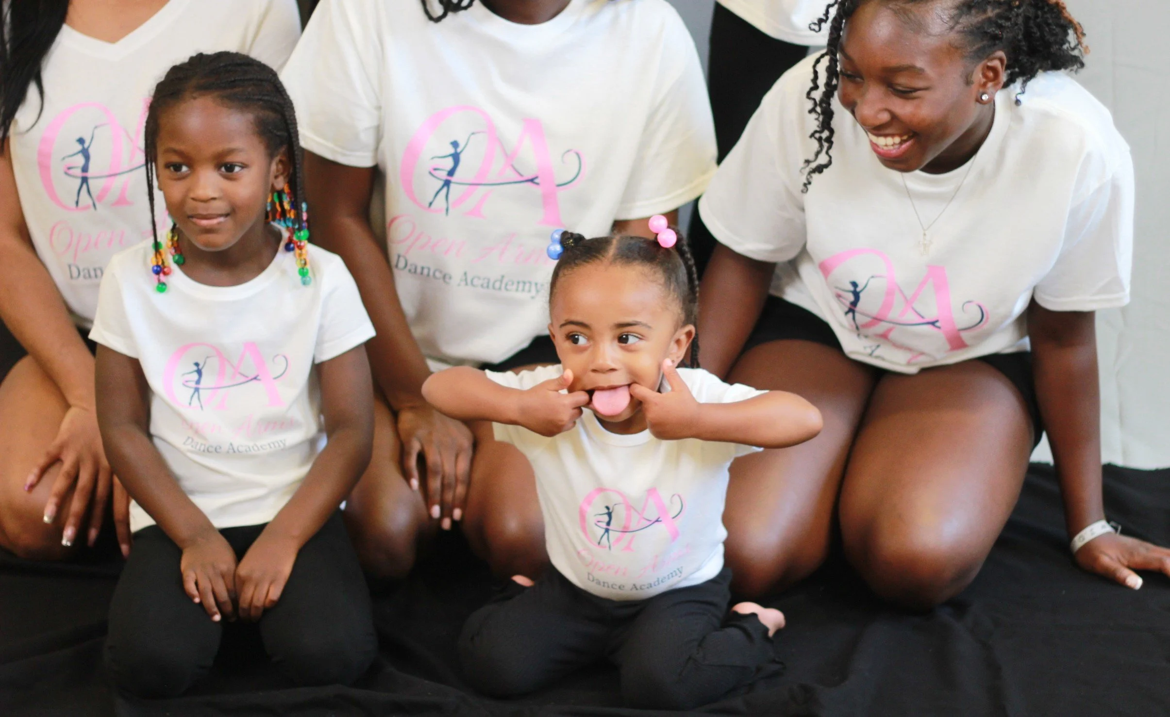 A group of young girls and women in matching dance academy t-shirts, some making playful faces, gathered sitting on a black surface.