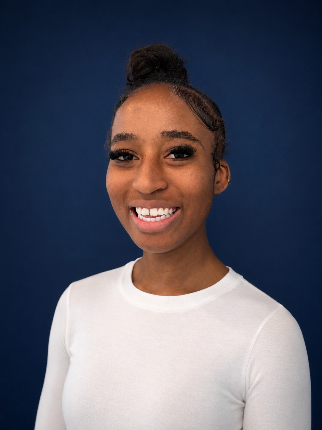 Close-up portrait of a young woman smiling, with dark curly hair styled in a bun, wearing a white top against a dark blue background.
