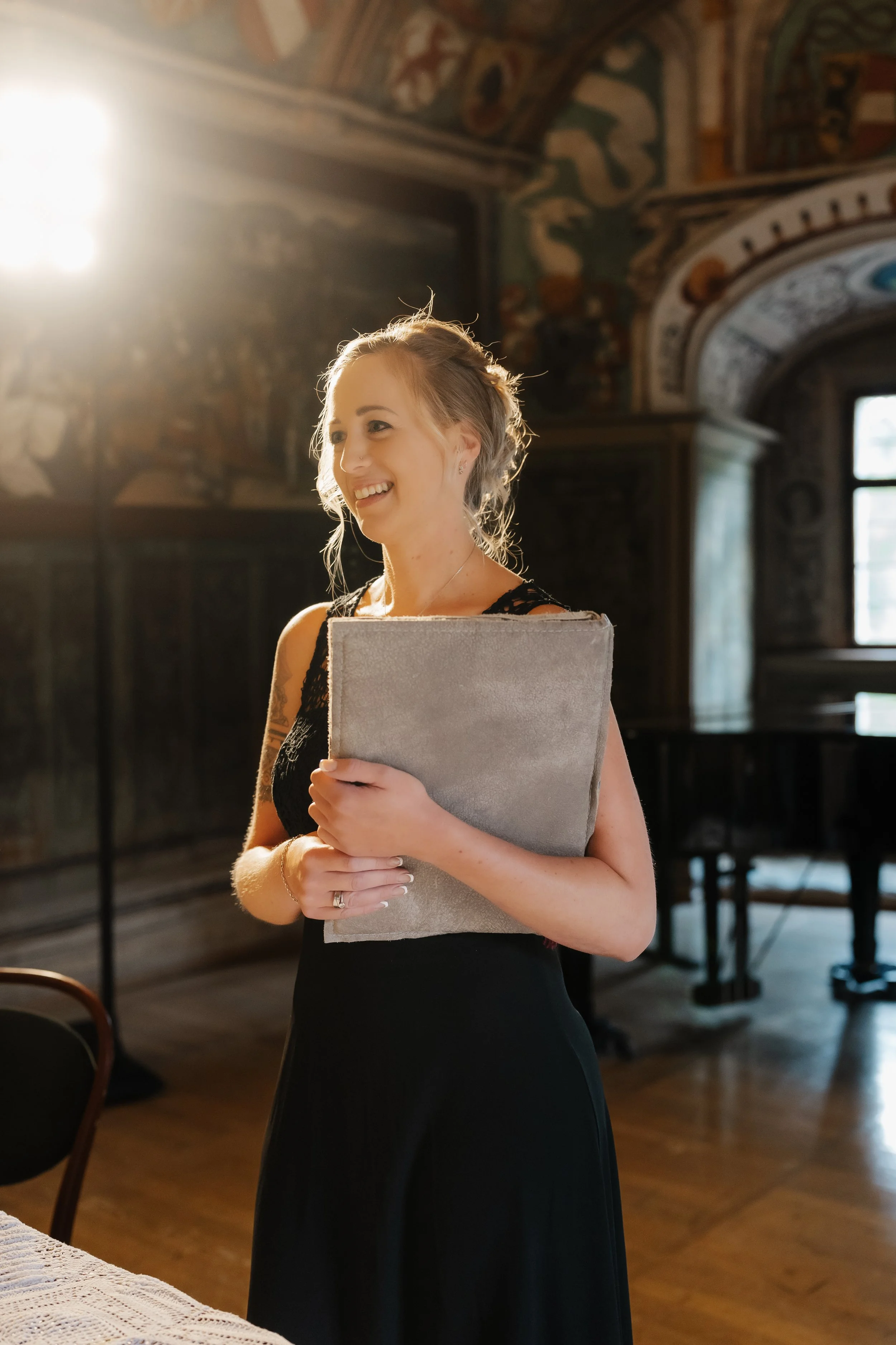 Eine lächelnde junge Frau trägt ein schwarzes Kleid und hält ein Buch in den Armen in einem historischen Raum mit Wandgemälden und einem Klavier im Hintergrund.