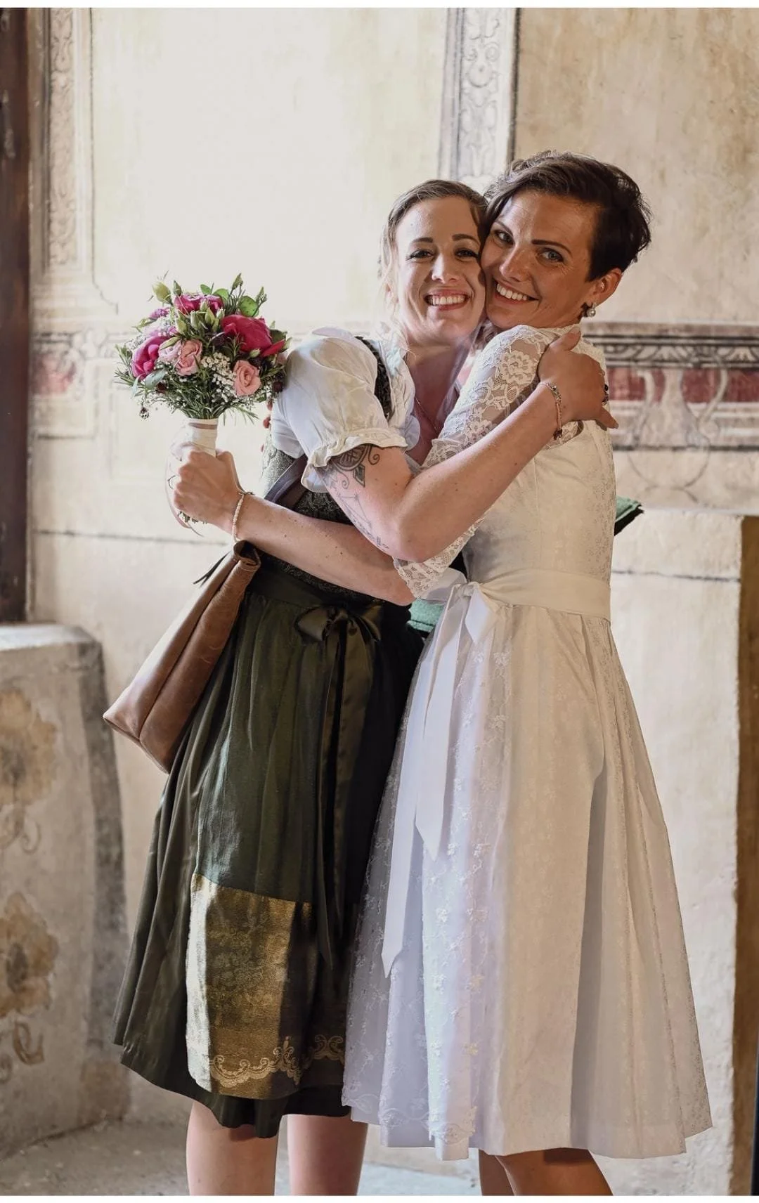 Zwei Frauen umarmen sich, eine trägt ein weißes Hochzeitskleid und die andere trägt traditionelle Trachtenkleidung, beide lachen bei einer Hochzeit.