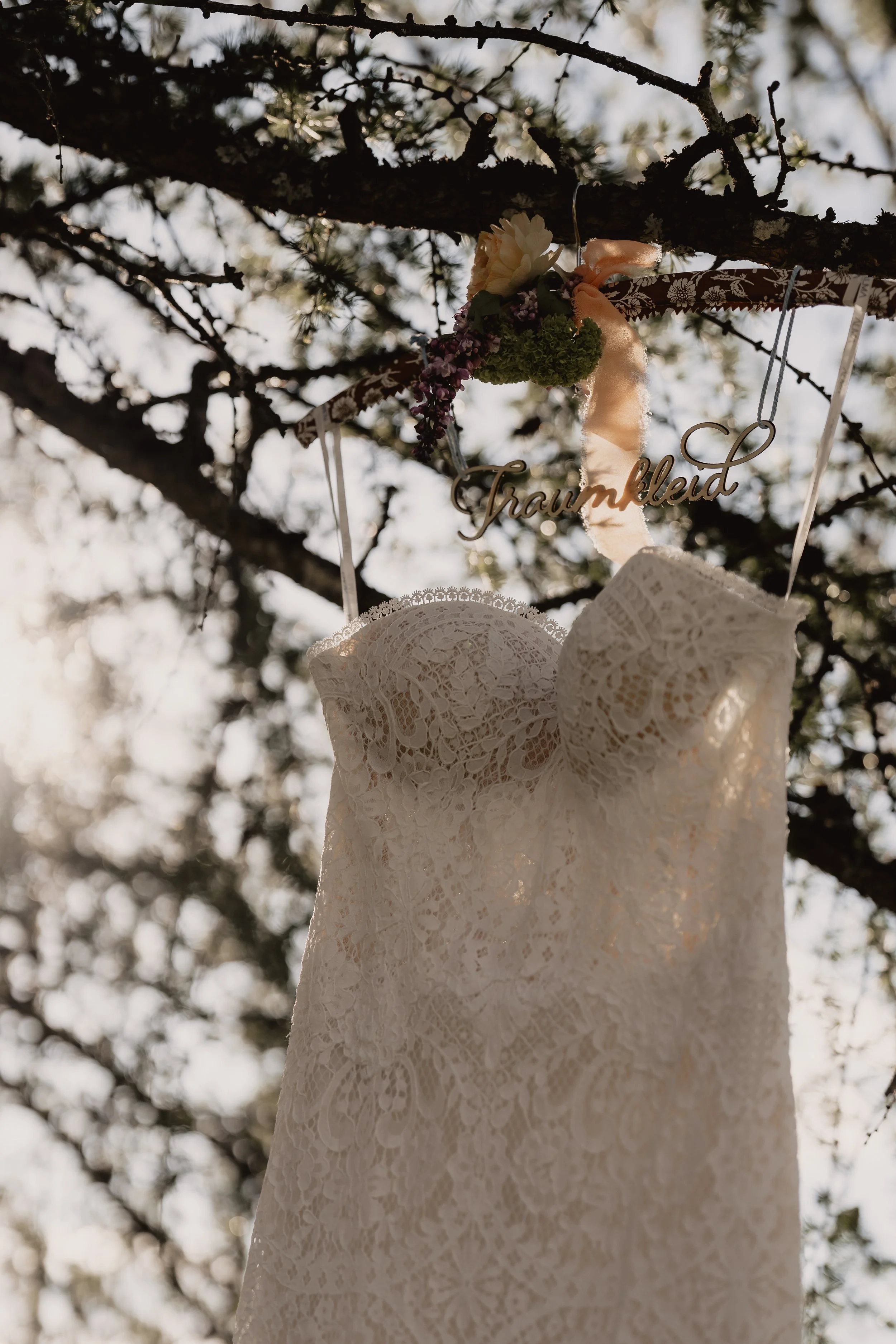 Hochzeitskleid aus Spitze, das an einem Baum im Freien hängt, mit einem dekorativen Namehrzschmuck und Blumen. Sonnenlicht durch die Blätter, romantische Atmosphäre.