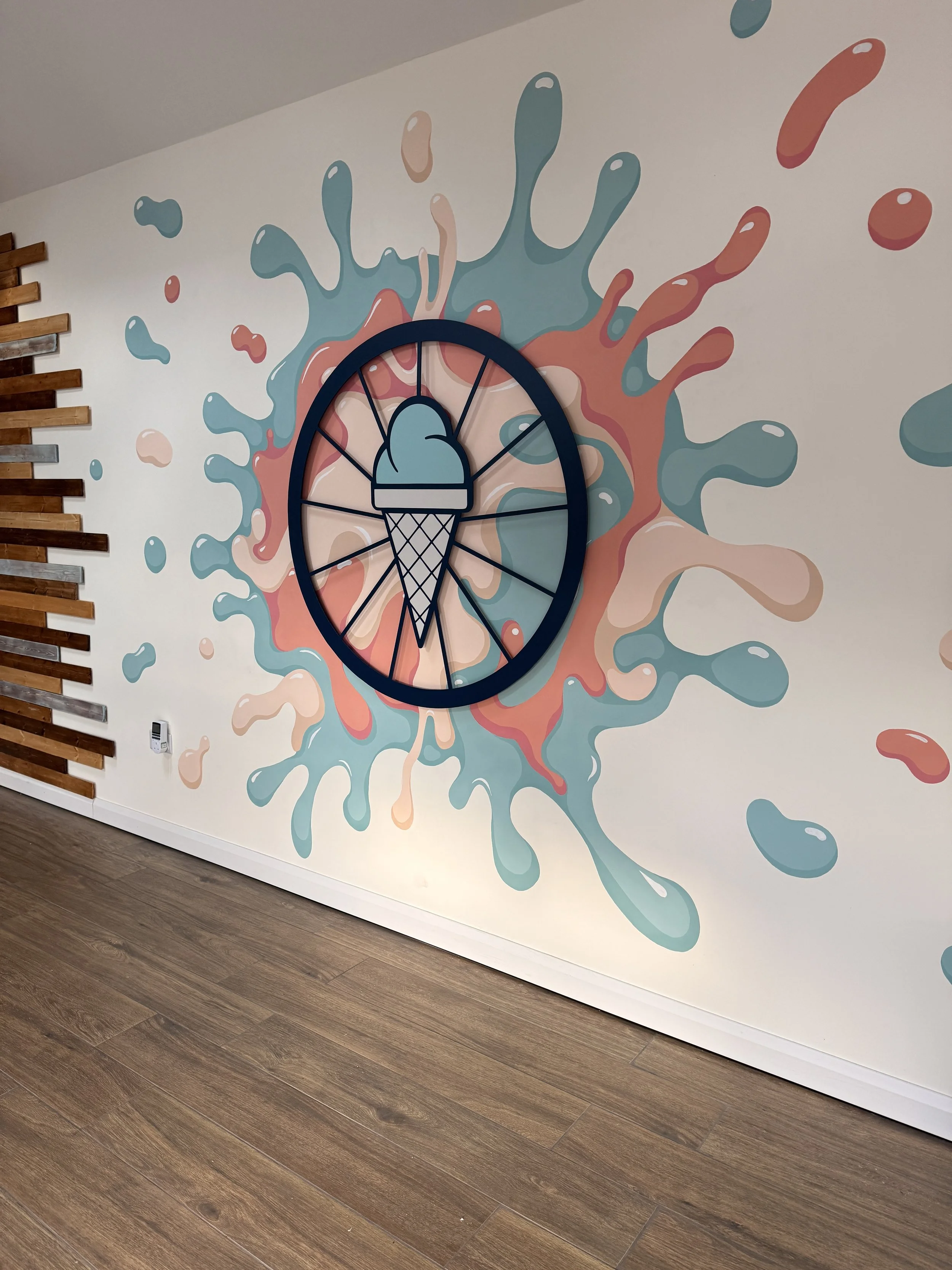 Decorative wall clock with a colorful splash background depicting an ice cream cone and abstract splashes in blue, peach, and orange, mounted on a wall with horizontal wooden slats and a wood floor.
