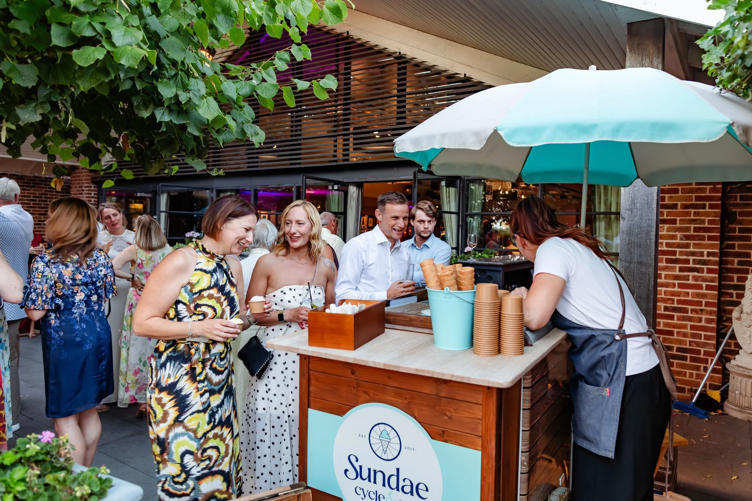 People socializing outside at a summer event, with a refreshment stand offering ice cream cones. An umbrella provides shade, and there are trees and brick building elements in the background.