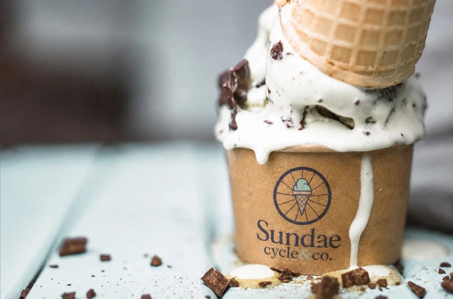 Close-up photo of a cup of ice cream with chocolate chunks and a waffle cone on top, in a paper cup with a 'Sundae Cycle & Co.' logo, on a light surface with chocolate pieces scattered around.