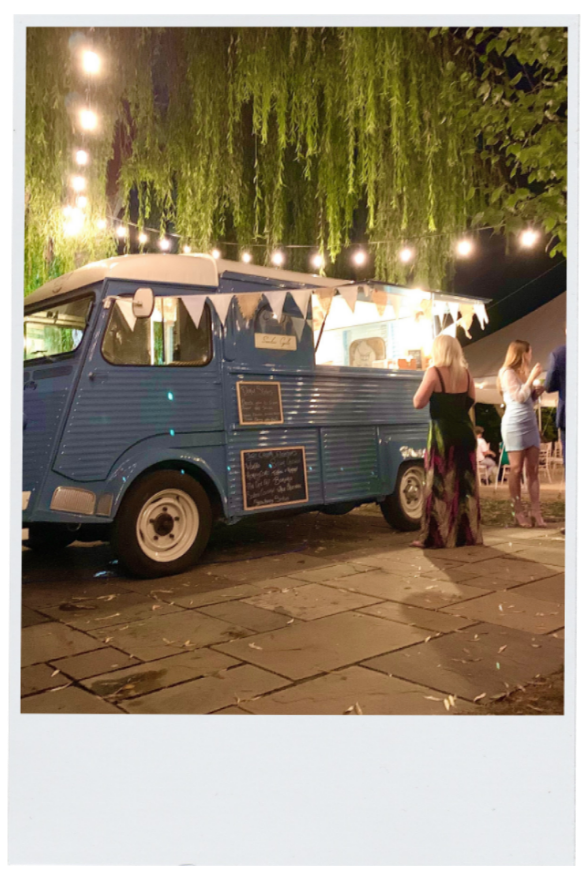 A vintage blue food truck decorated with string lights and bunting, parked on a stone-paved area at night with people nearby, under hanging trees.