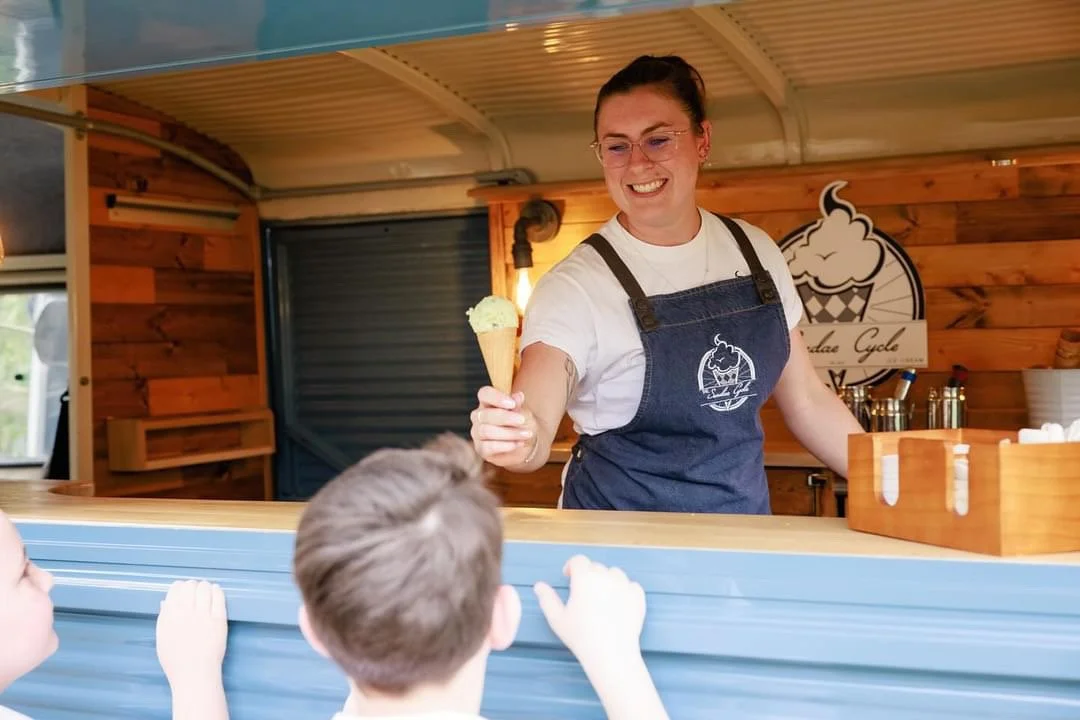 A woman serving ice cream cone to children at an ice cream stand inside a food truck.