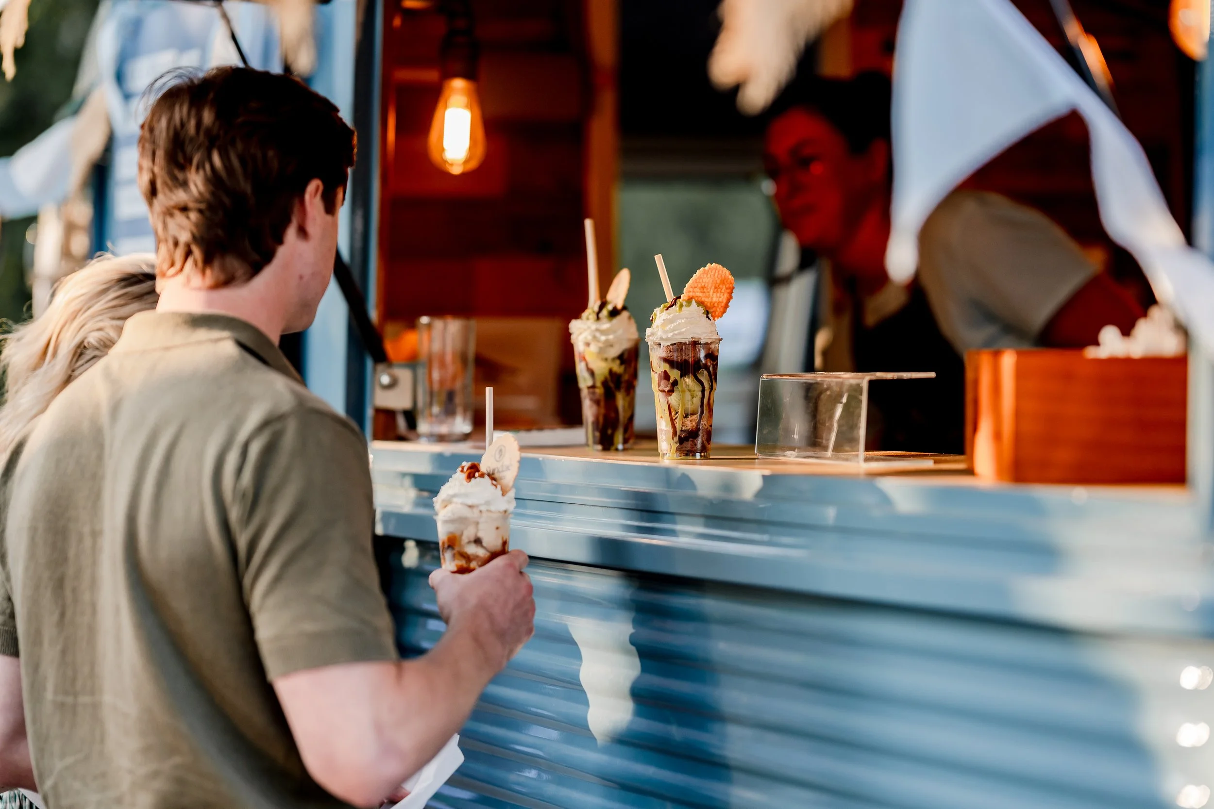 A person holding a dessert cone with whipped cream and chocolate syrup, standing at a food truck serving milkshakes topped with whipped cream, chocolate sauce, and a waffle cone piece in warm lighting.