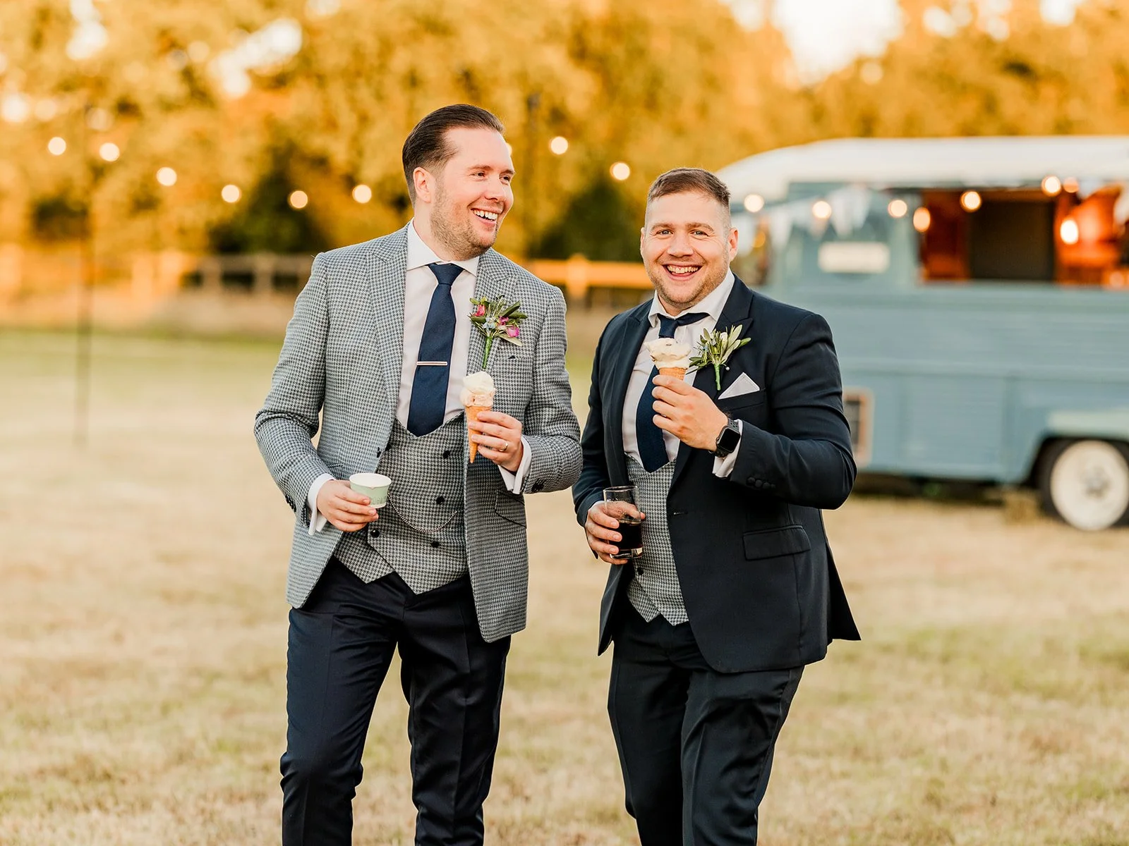 Two men in formal suits enjoying ice cream cones and drinks outdoors during the fall, with a vintage trailer and autumn trees in the background.