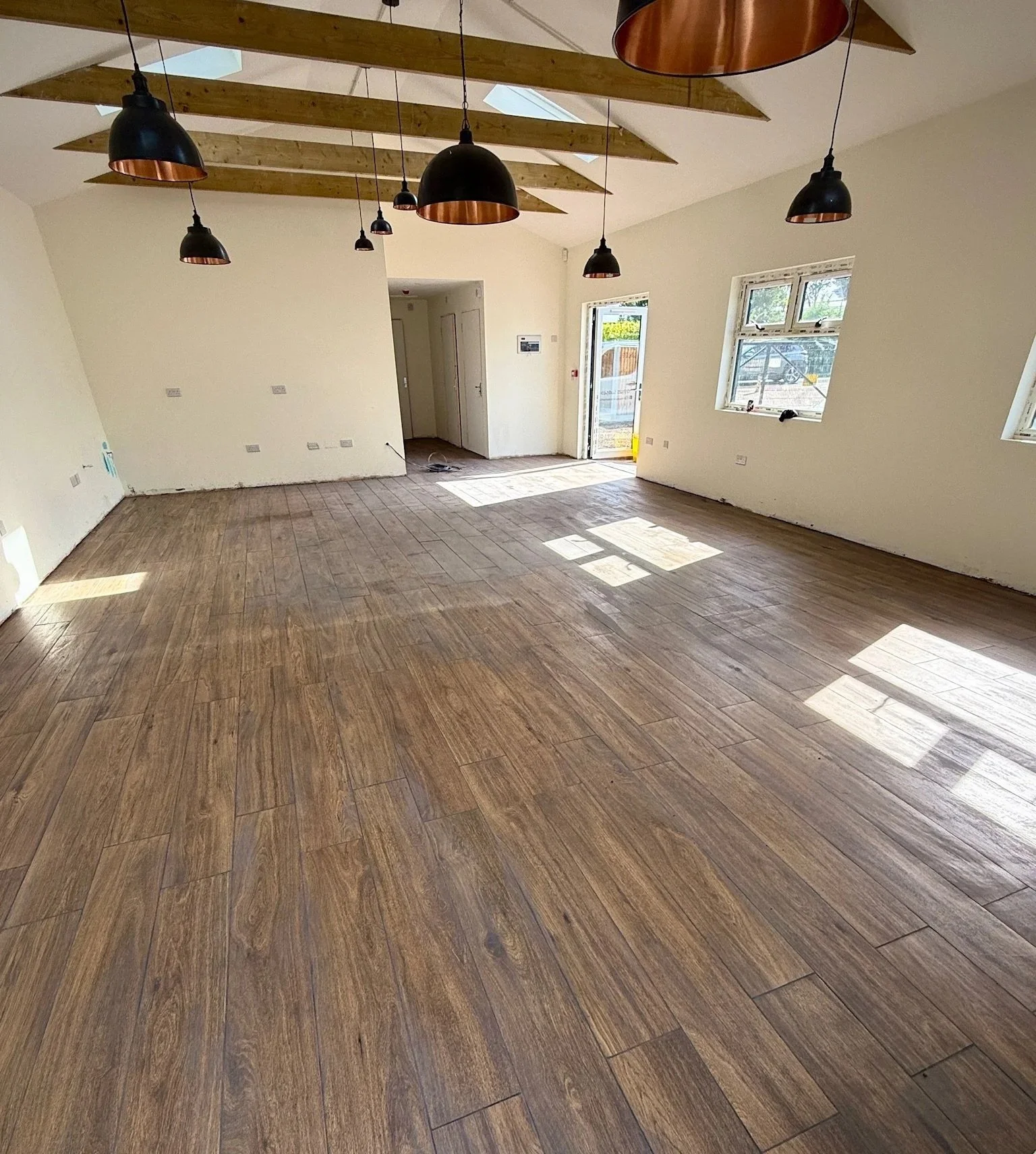 Empty room with wood flooring, white walls, several windows, a door, and hanging black and copper pendant lights.