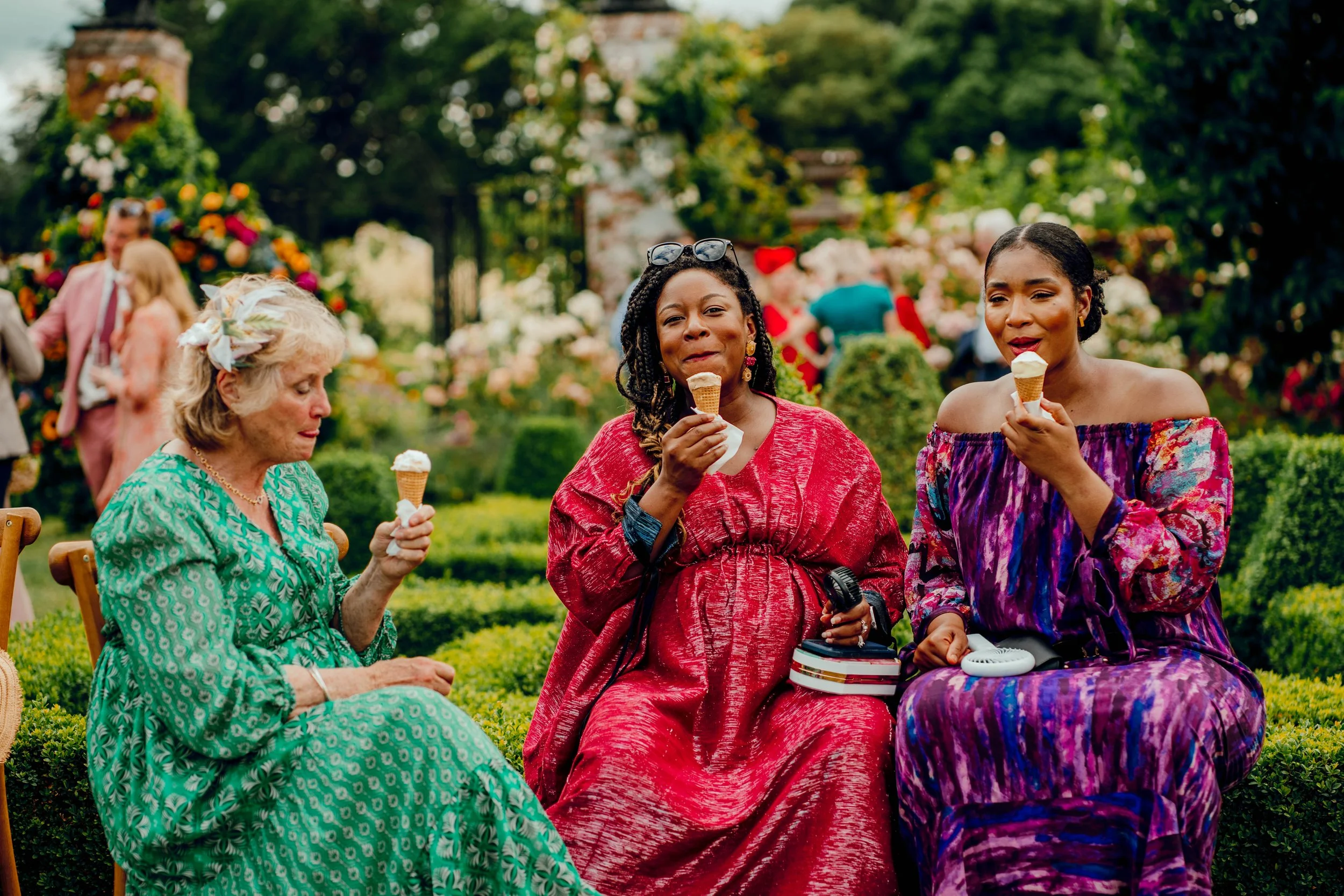Three women sitting outdoors enjoying ice cream cones, with two women in patterned dresses and the woman in the middle wearing red and taking a selfie. In the background, other people are socializing near decorative floral displays.