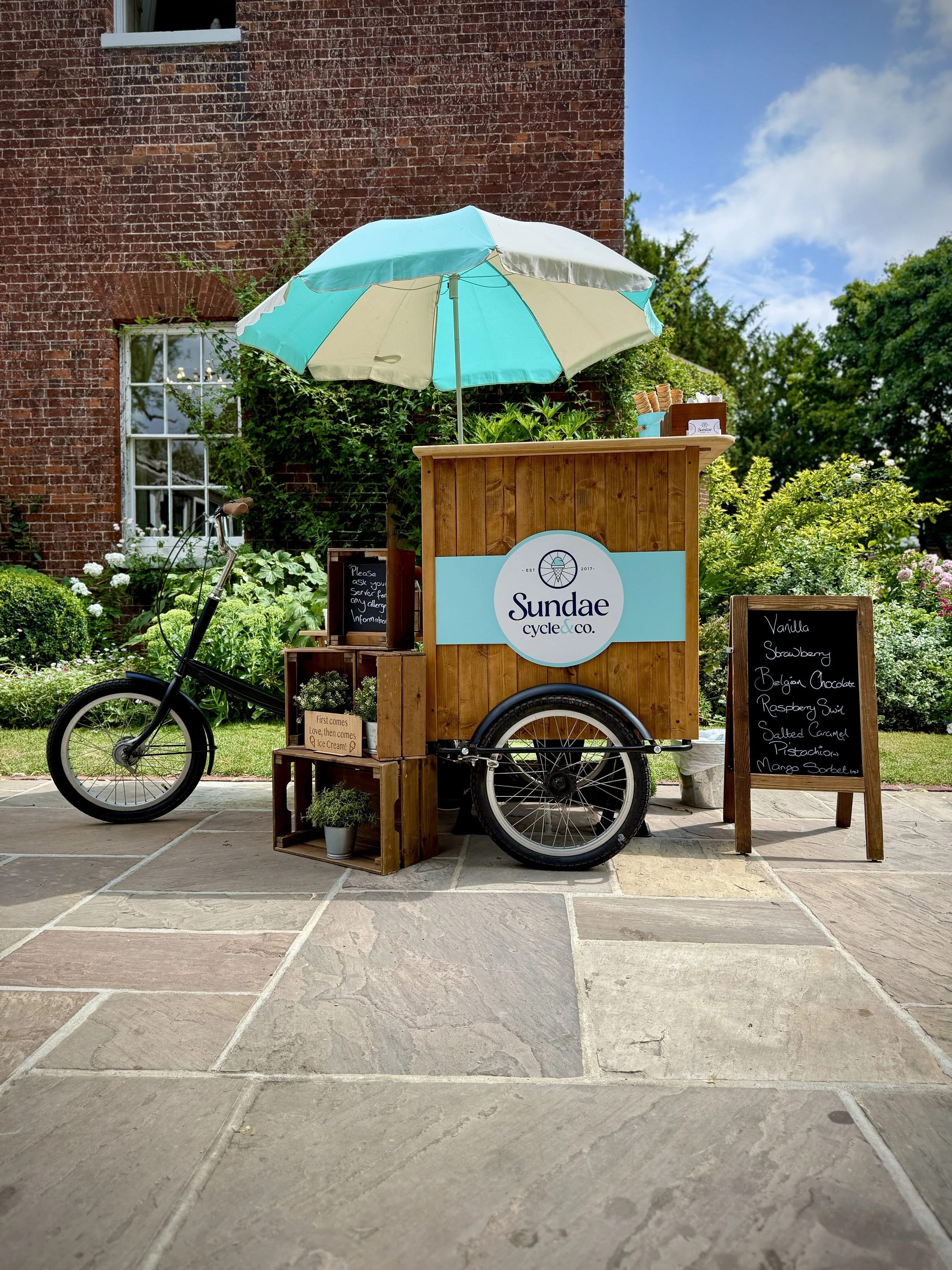A bicycle cart with a large blue and white umbrella, advertising Sundae cycle & co., is parked on a patio with tan and gray tiles. The cart has a wooden front panel with the logo, a small chalkboard sign listing ice cream flavors, and a small potted plant. In the background, there is a brick building, a window, and greenery with trees and bushes under a partly cloudy blue sky.