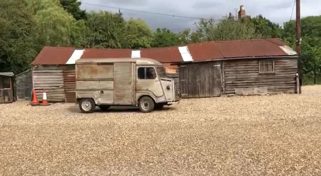 An old wooden building with a rusted metal roof, a weathered van parked in front, and orange traffic cones nearby.