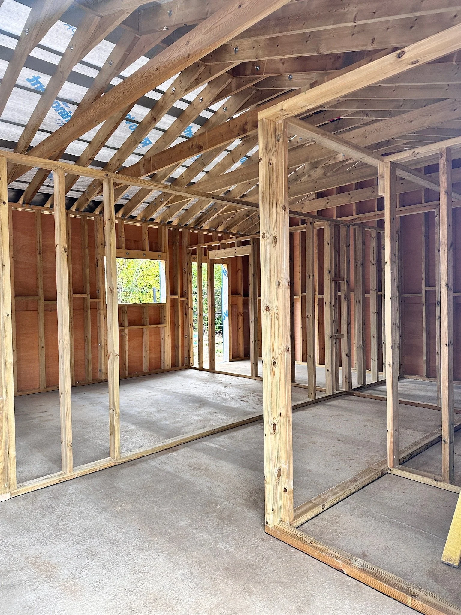 Interior view of a house under construction showing wooden framing and roof trusses, with a concrete floor and an open window.