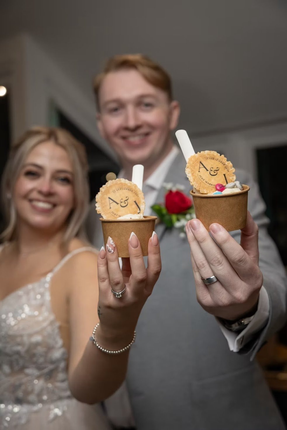 A smiling bride and groom holding matching cups of ice cream with cookies that have capital letters A and J, possibly at their wedding celebration.