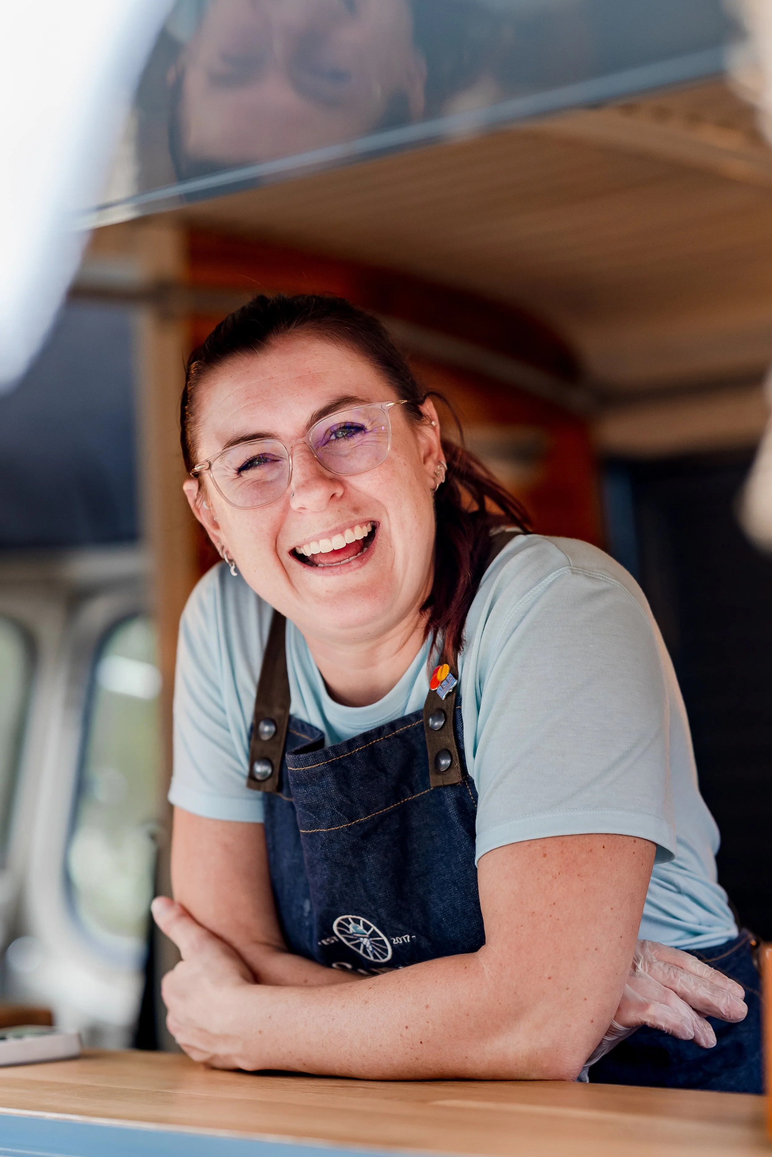A woman with glasses and earrings leaning on a counter, smiling and looking at the camera, wearing a light blue shirt and a denim apron, with a wooden ceiling and window in the background.