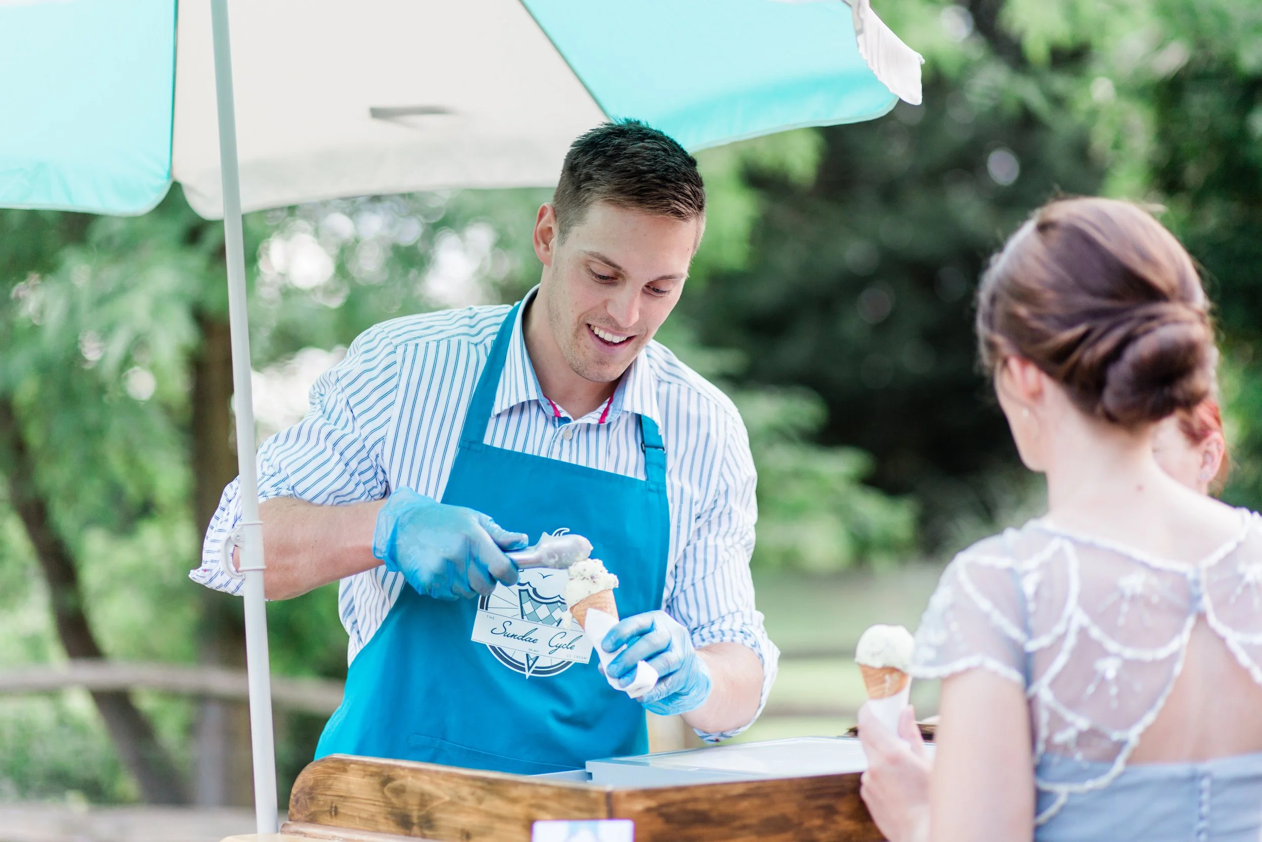A man in a blue apron and striped shirt serving ice cream to a woman in a white blouse at an outdoor ice cream stand, with a green leafy background and a white and green umbrella.