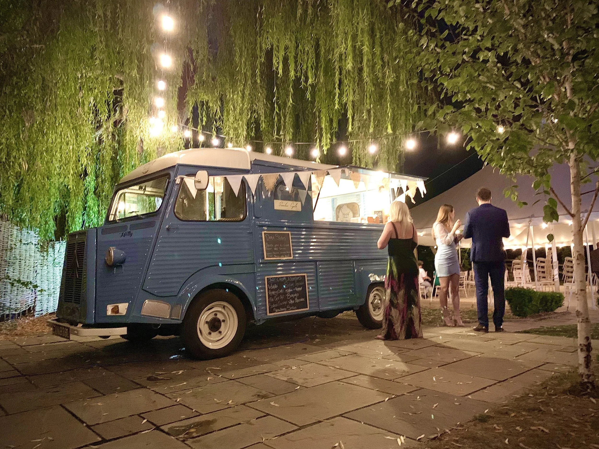 Night scene of a food truck with bunting decorations, people standing nearby, and trees illuminated with string lights.