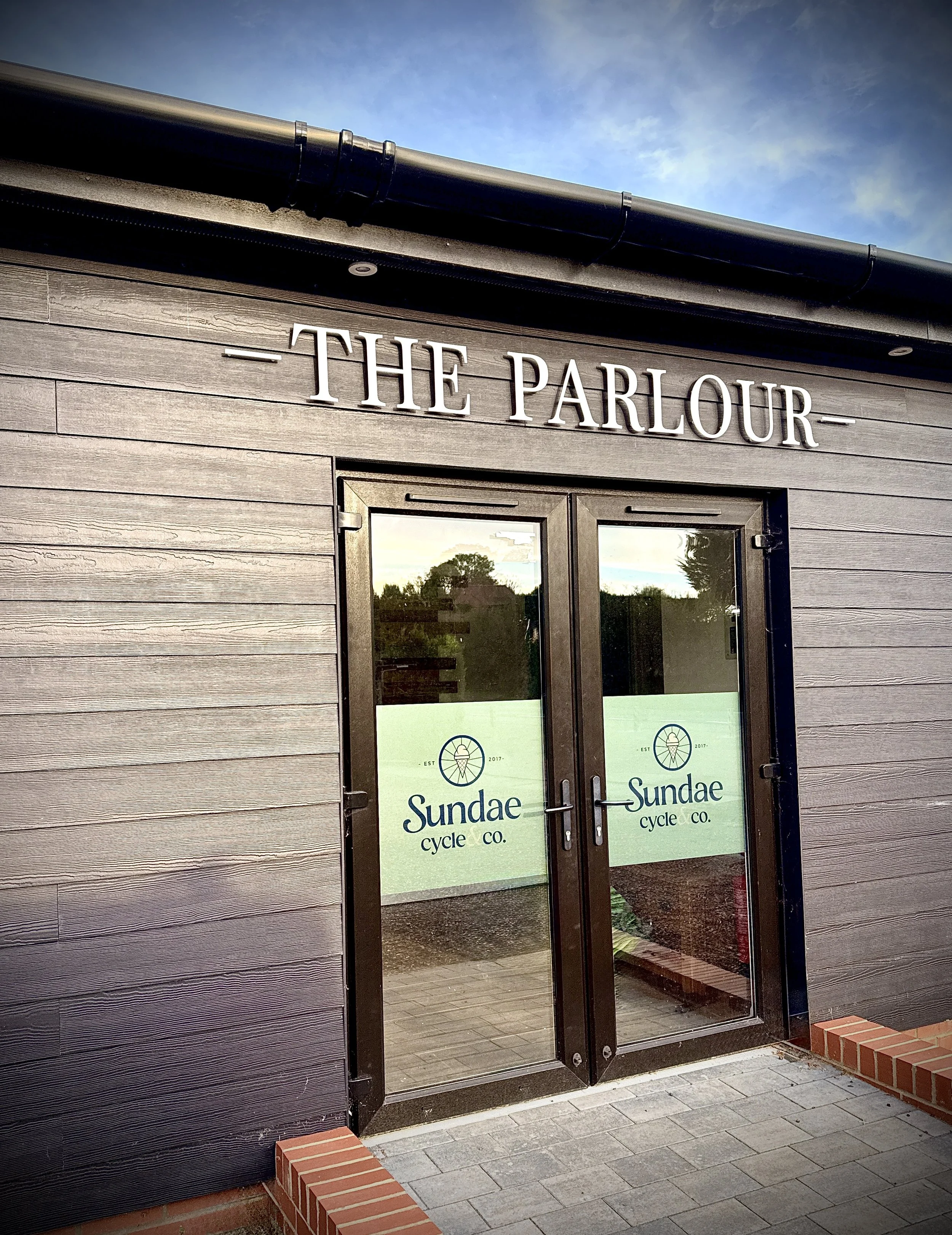 The exterior of a building with a sign that reads "The Parlour" above a door. The door has glass panes with a sign that says "Sundae cycle co." The building is made of horizontal wooden planks, and there are small brick steps at the entrance. The sky is partly cloudy with blue visible.