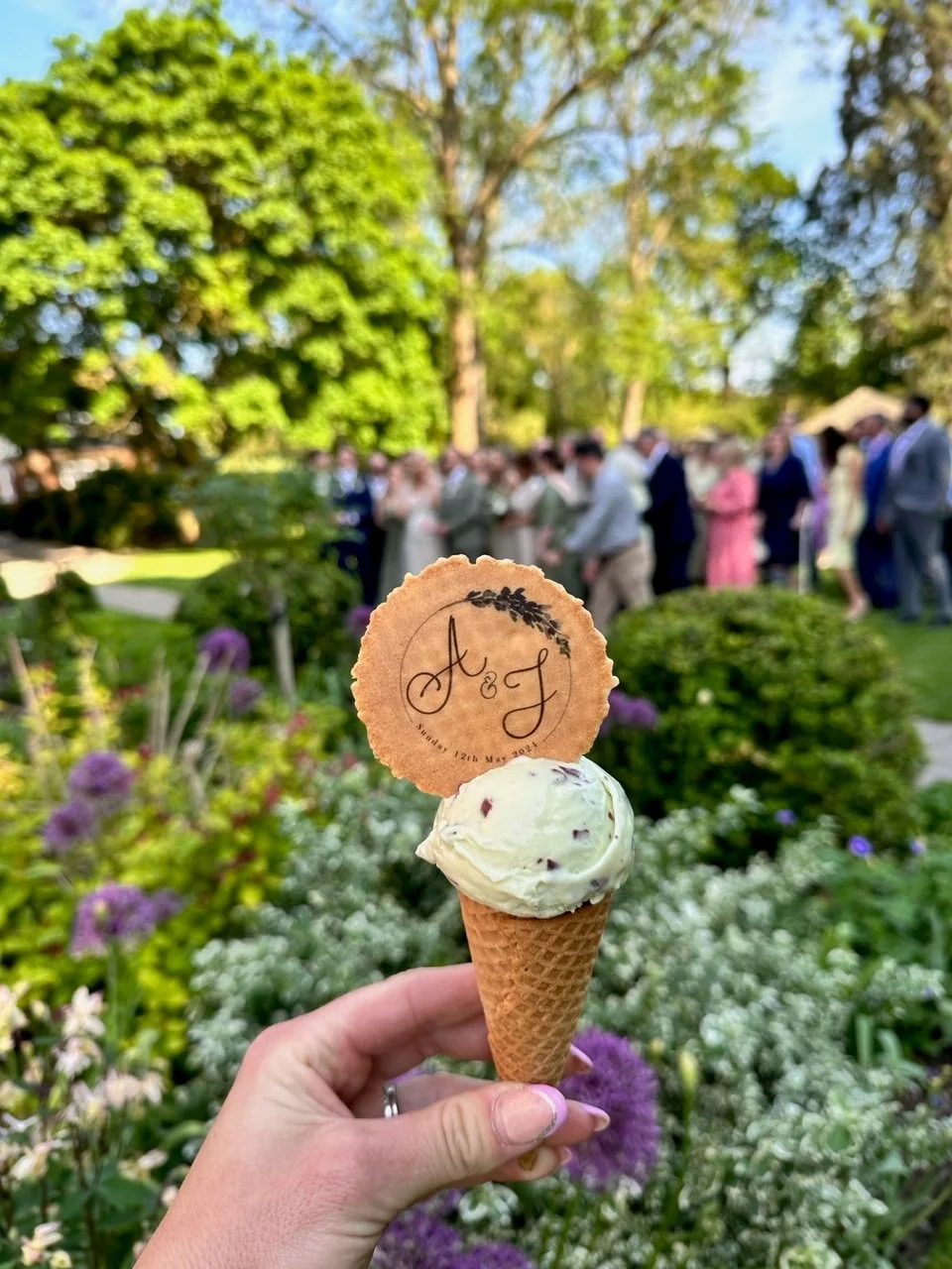 Hand holding an ice cream cone with a decorative cookie on top, engraved with the initials 'A & J' and a date, at an outdoor wedding gathering with greenery and guests in the background.