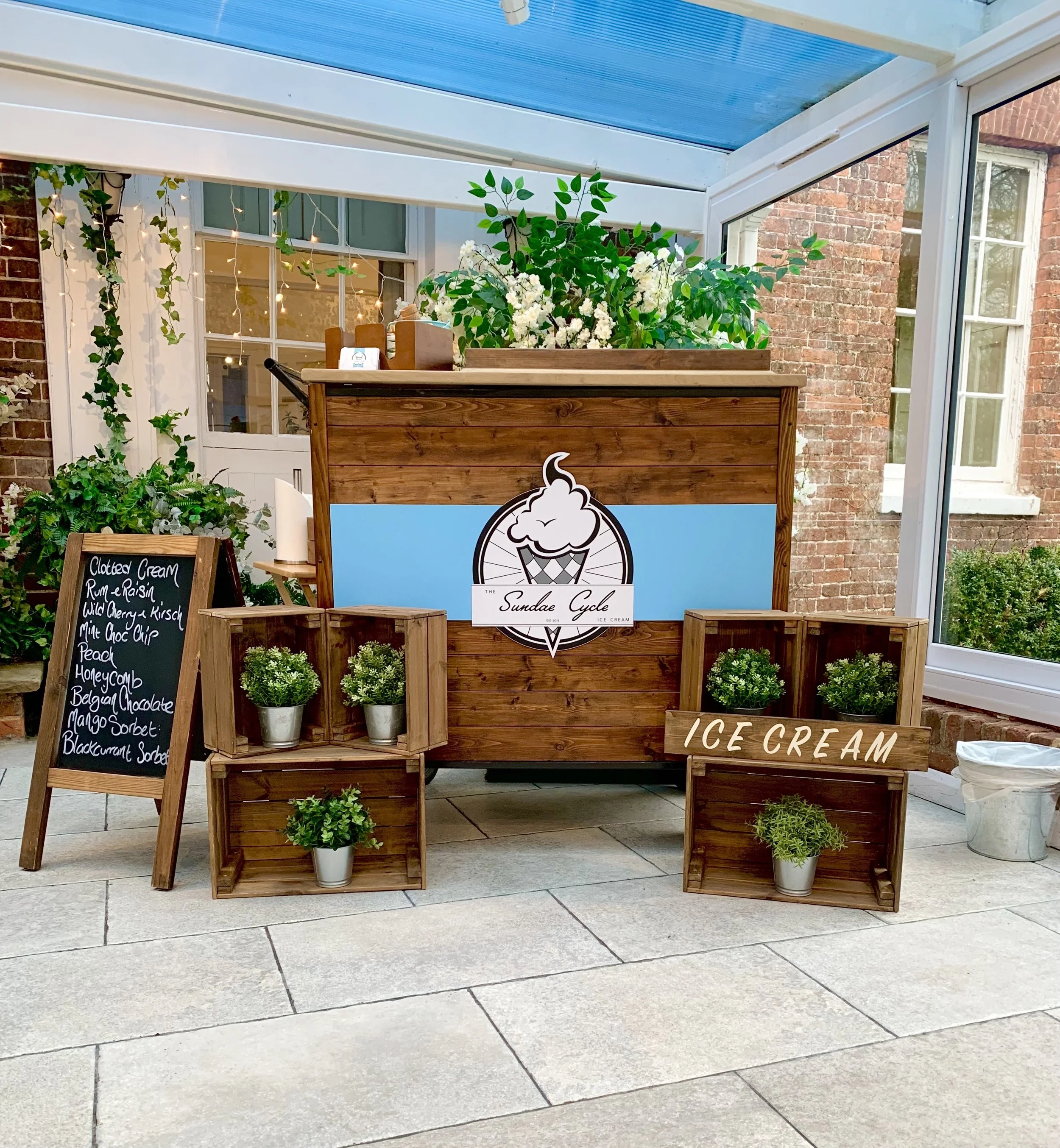 Wooden ice cream stand with a large sign displaying a cone and the words "The Sundae Cycle." There are green plants in pots on and around the stand, and a black chalkboard menu listing various ice cream flavors next to the stand. The background shows a brick building and a glass-enclosed porch with string lights and green vines.