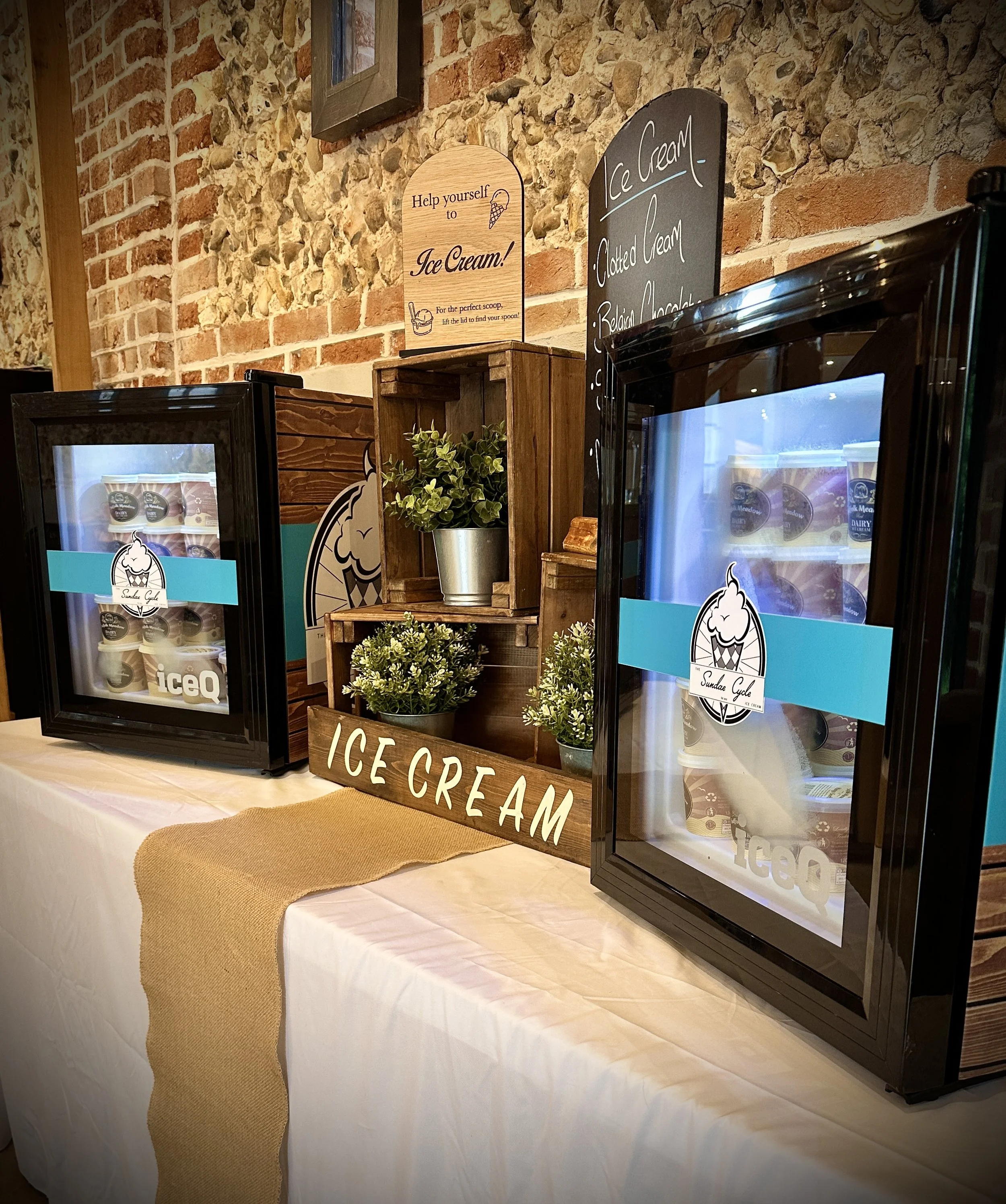 Display of ice cream containers on a table with a decorative sign reading 'Help yourself to Ice Cream' and a blackboard menu listing ice cream flavors, with plants and a brick wall in the background.