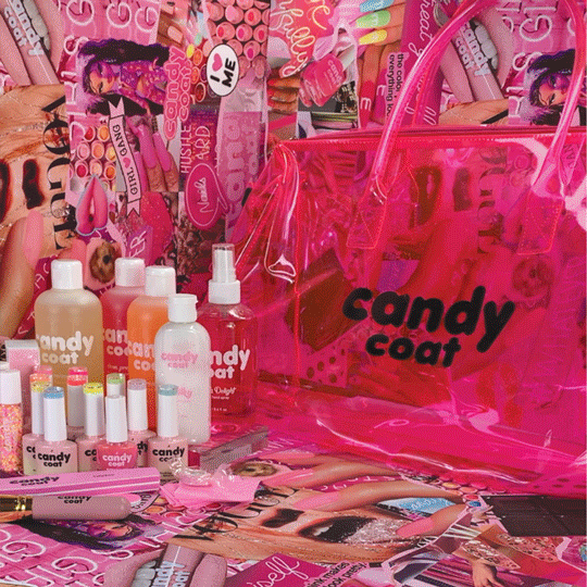 A collection of pink and white Candy Coat nail polish bottles, lip glosses, lipsticks, beauty accessories, and a large pink transparent bag with the Candy Coat logo, all arranged on a table with a background of pink and purple makeup and beauty product packaging.