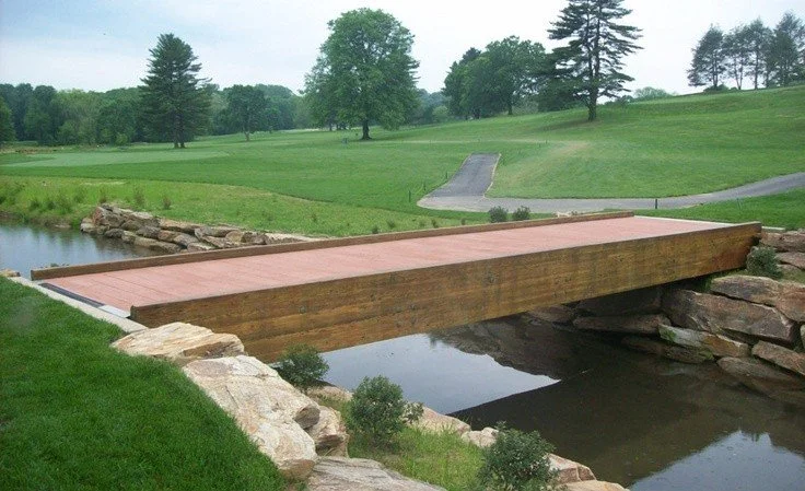 A small wooden footbridge over a creek in a park or golf course, with a grassy landscape and trees in the background.